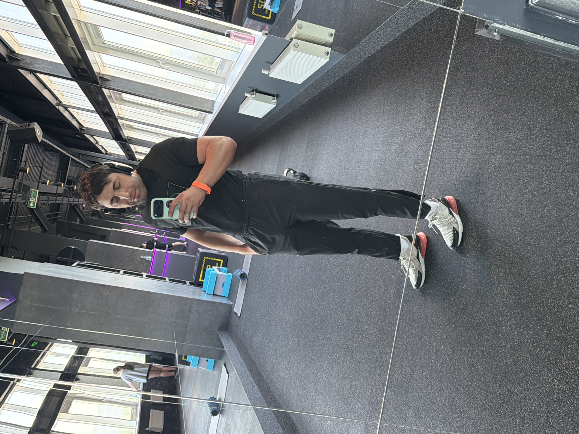 The image depicts a modern fitness center located in Krasnoyarsk, Russia. A male individual, appearing to be in his 30s, stands in the foreground, taking a mirror selfie with a light green smartphone held in his right hand. He wears black over-ear headphones, a black short-sleeved t-shirt, black athletic pants with zippered ankles, and light-colored athletic shoes with red/orange accents. An orange fitness tracker or band is present on his left wrist, and his left hand is in his pants pocket.
The fitness center features dark gray, textured rubberized flooring and large mirrored walls on the left, reflecting the interior. A series of tall, rectangular windows line the right side of the room, offering an outdoor view of distant buildings and sky, indicative of an elevated floor. The ceiling is dark, exposing structural elements, ductwork, speakers, and overhead lighting. A green "ВЫХОД" (Exit) sign in Cyrillic script is visible on the ceiling.
Reflected in the mirrors are several objects and other individuals:
On the left, a woman wearing a light gray t-shirt and dark shorts stands facing away from the camera, near the windows.
On the right, a man in a dark t-shirt and shorts is partially visible in the reflection near a dark counter.
Various gym equipment is present, including blue step platforms, a black and yellow exercise box with a digital timer displaying "50," gray and black yoga mats, and a black foam roller.
Below the windows, white radiators are mounted on a dark wall. A clear water bottle and a pink water bottle are visible on the window ledge or a surface below. A dark kettlebell is partially reflected on the floor near the main subject's feet. Purple neon strip lighting accents parts of the gym's structure.
