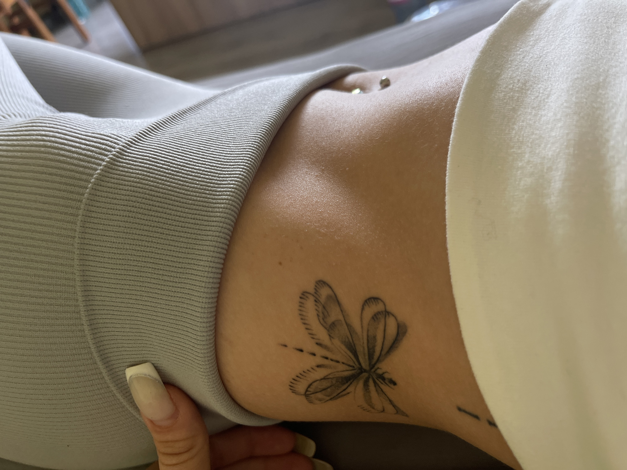 A close-up, low-angle shot shows the side of a person's torso. The person is wearing light gray, ribbed athletic wear and a white garment draped over their shoulder. A dragonfly tattoo is visible on their lower abdomen. A navel piercing is also present. A fingernail is in the lower left corner of the frame. The background is out of focus and appears to be a domestic setting.