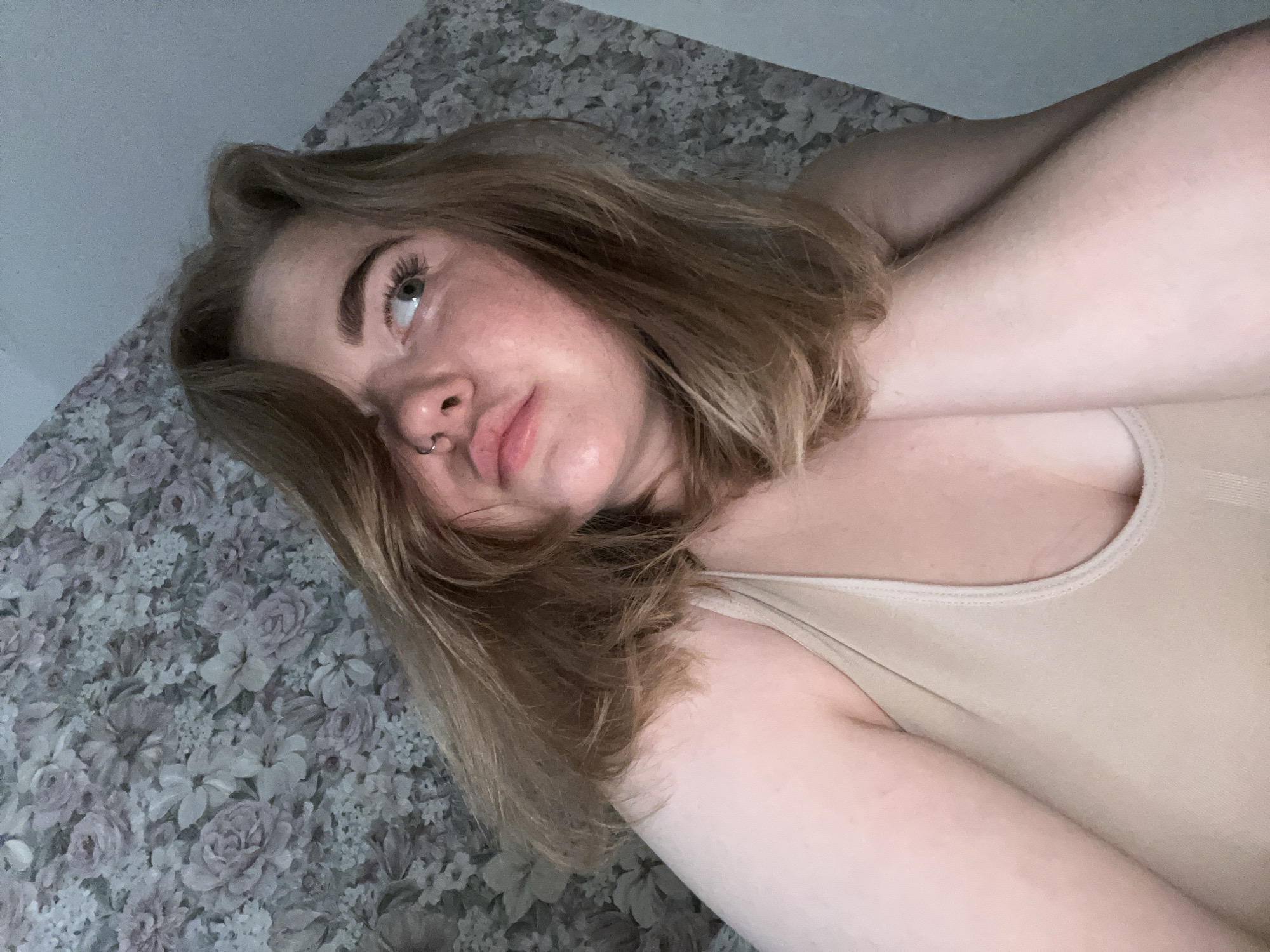 A woman with fair skin and short blonde hair is lying down. She is wearing a light beige tank top. Her arms are crossed and raised behind her head. She has a nose ring in her left nostril. She appears to be lying on a bed with a floral patterned bedspread. The location is not identifiable as Chaikovsky, Russia.