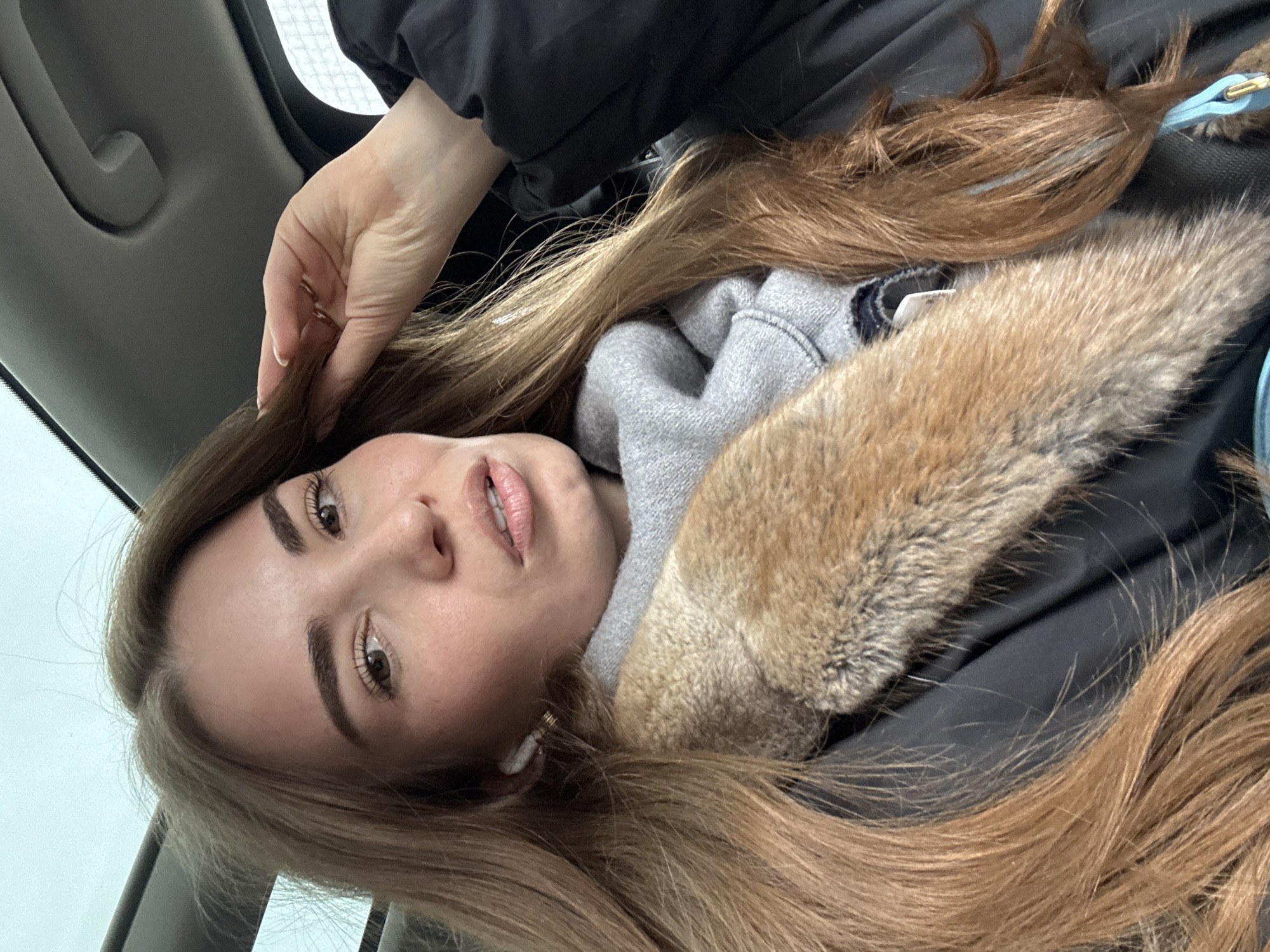 A woman with long, light brown hair is in a vehicle. She is wearing a black jacket with a gray hooded sweatshirt and a fur-lined hood. Her left hand is near her hair. She has an earbud in her left ear. The setting suggests the interior of a car, based on the interior panel visible in the background. The location in Moscow, Russia, cannot be determined from the visual information provided.