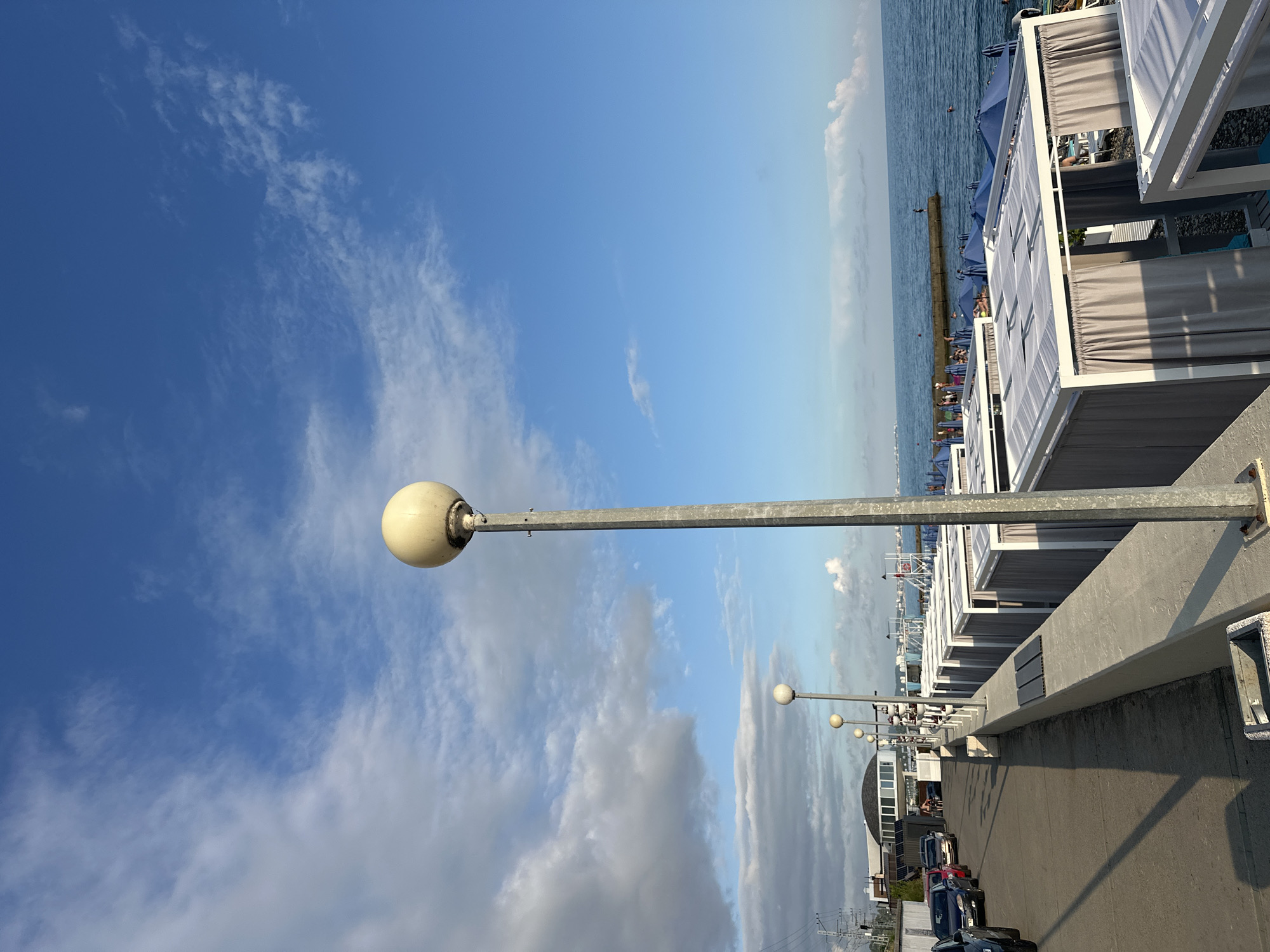 This is a vertical, eye-level, outdoor shot of a beachside promenade. The sky is a vibrant blue with scattered wispy white clouds, suggesting a clear day. The sun is casting long shadows, indicating it is likely late afternoon or early evening.
The scene features a row of beach cabanas with white frames and grey curtains, facing the sea. In the foreground, a tall, metal lamppost with a round, cream-colored light fixture stands prominently against the sky. Further down the promenade, more lampposts of a similar design are visible, also stretching towards the horizon.
The sea is a deep blue and appears calm, with a few people visible in the water and along the shoreline. There is a concrete barrier or pier separating the promenade from the sea. Along the promenade, a few cars are parked, including a dark blue sedan, a red car, and other vehicles. A building with a domed roof is also visible in the background, along with what appear to be power lines.
The overall mood is peaceful and relaxed, characteristic of a seaside resort. The location appears to be Krayevsko-Armyanskoye, Russia. No visible text is present in the image.