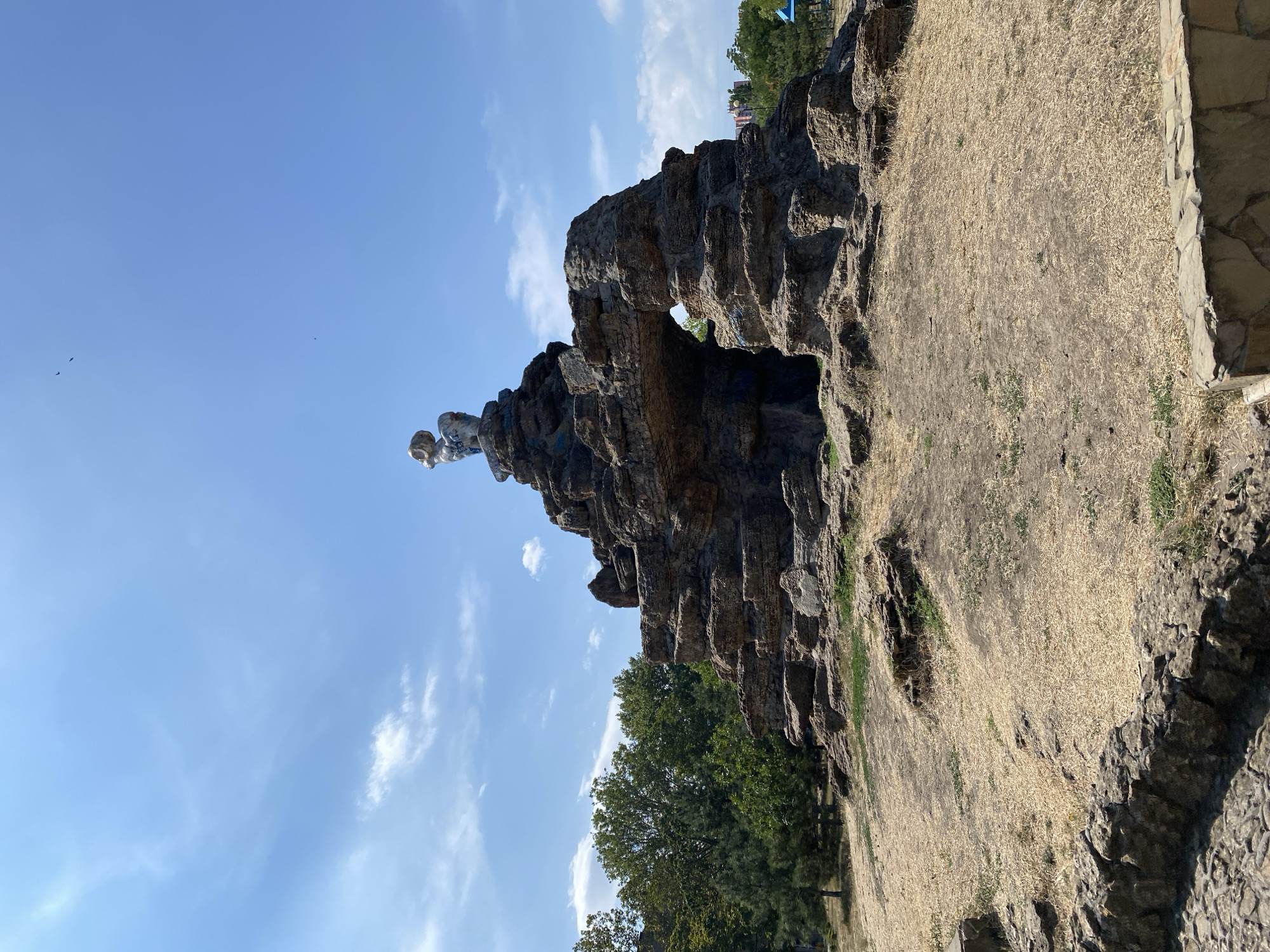 The image depicts a dry, grassy hill with a man-made rock formation in the center. The rock formation resembles a small mountain with an arched opening. Atop the highest point of the rock formation sits a statue of a ram. The surrounding area is sparse with dried grass and some scattered rocks. In the background, there are trees and buildings, suggesting an urban or park setting. The sky is blue with scattered white clouds. This landmark is identified as the "Ram" or "Aries" sculpture in Chornomorsk, Ukraine, located in the city's central park.