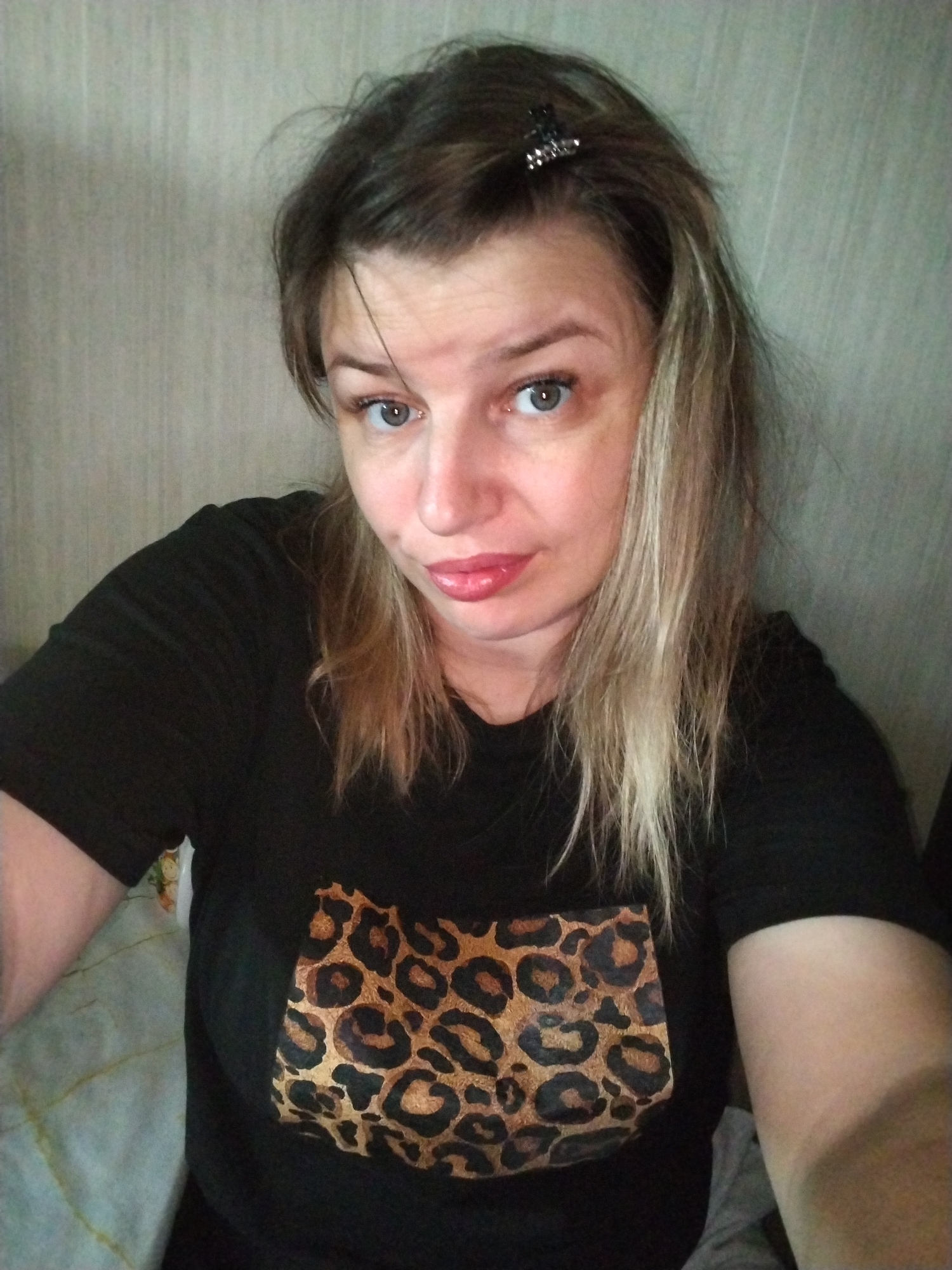 A young woman with blonde-streaked brown hair, wearing a black t-shirt with a leopard print design on the chest, is captured in a medium shot against a textured light-colored wall. She has blue eyes and is wearing lip gloss. A dark hair clip is visible in her hair. The image is an indoor, eye-level selfie, with the subject positioned slightly off-center to the right. The background appears to be domestic, suggesting a home environment. No specific location in Yaroslavl, Russia is identifiable from the visual cues.
