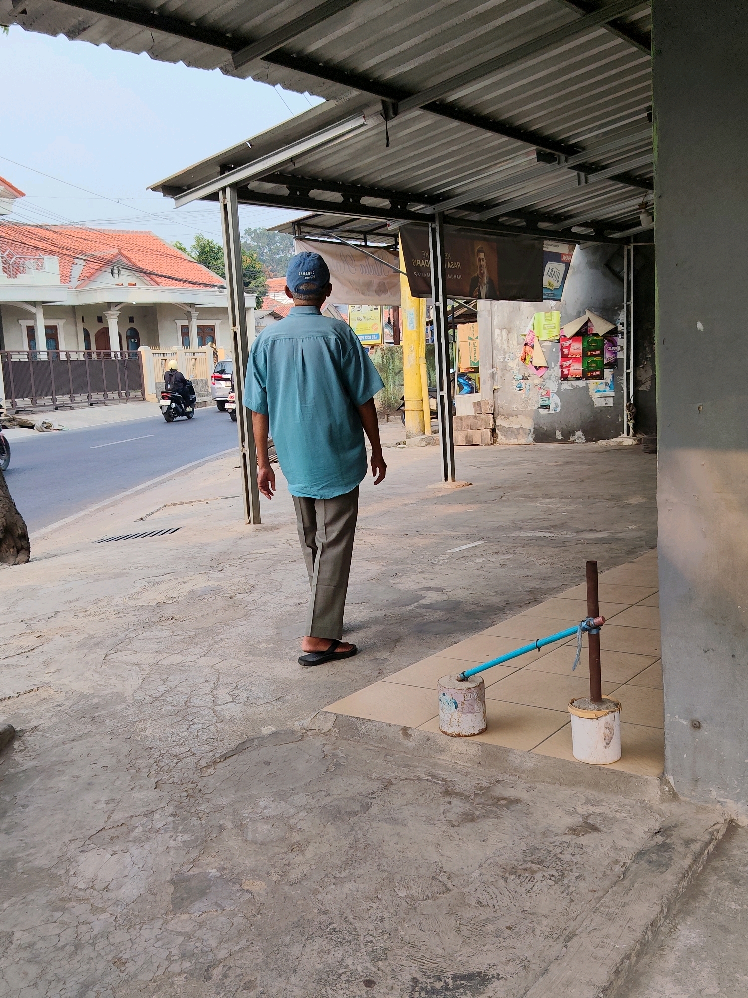 In Purwakarta, Indonesia, an elderly man wearing a teal short-sleeved button-up shirt, grey trousers, and black flip-flops walks away from the camera down a sidewalk. He wears a blue baseball cap. A corrugated metal roof supported by metal posts extends over the sidewalk. A street runs parallel to the sidewalk. Two motorcycles and a car are visible on the street. On the sidewalk, a makeshift water dispensing system consists of a blue pipe connected to a spigot, supported by two rusted cans. The environment appears to be a commercial or residential area with buildings visible in the background.