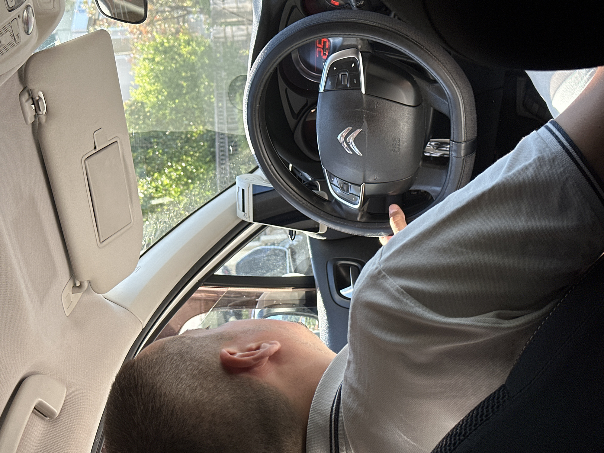 An individual, from the shoulders up, is seated in the driver's seat of a Citroën car, facing forward. The person has short, buzzed brown hair and is wearing a light gray polo shirt with dark stripes on the collar. Their left hand is visible on the steering wheel, which is covered with a black steering wheel cover. The car's dashboard shows a digital display with the number "25" illuminated in red. A small, white electronic device is mounted on the dashboard to the left of the steering wheel. Outside the car, through the windshield and side windows, lush green trees and foliage are visible, suggesting an outdoor environment, likely in Budva, Montenegro, given the context. Sunlight streams into the car, creating highlights and shadows. The interior of the car appears to be a typical passenger vehicle.