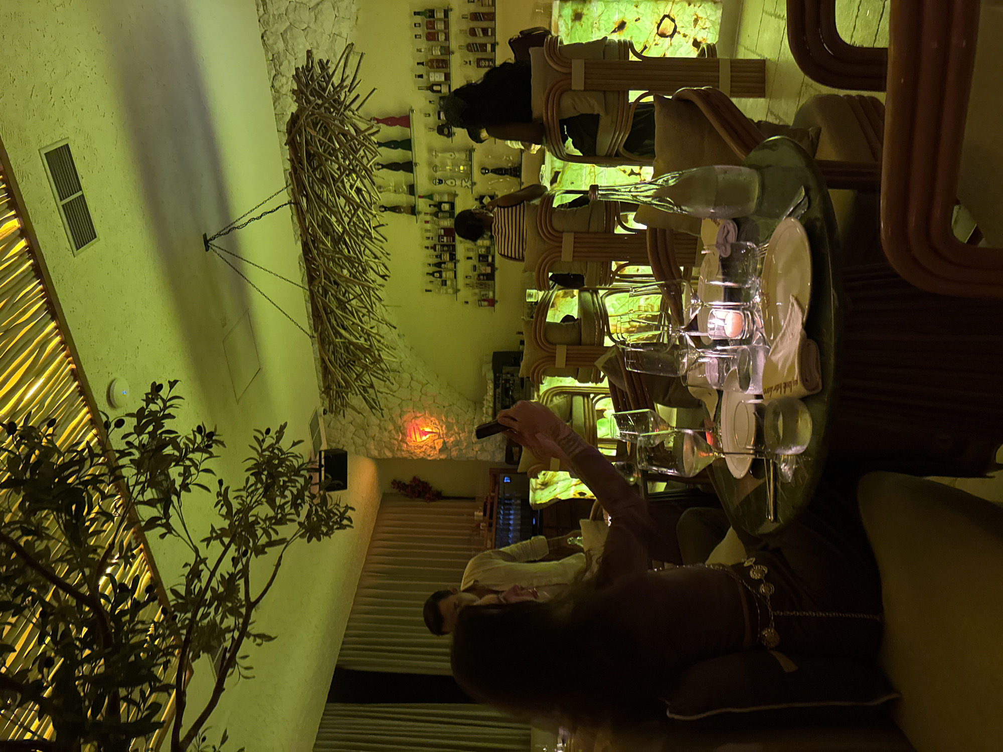 The image depicts the interior of a restaurant in North Kuta, Indonesia, illuminated by a green-yellow ambient light. In the foreground, a woman is seated at a round table, holding up a smartphone to take a photo. The table is set with place settings, including white plates, wine glasses, a water glass, a lit candle in a small glass holder, and a bottle of water. The woman is wearing a brown top and a decorative chain belt. To her left, another person is partially visible, seated. Behind her, a man in a light-colored shirt stands, looking towards the bar area. The restaurant features a bar in the background, with shelves stocked with numerous bottles of alcohol and decorative items. Bar stools with wicker frames and cushioned seats are arranged along the bar. The walls appear to be made of rough stone, and a large decorative light fixture made of branches hangs from the ceiling. Artificial foliage, resembling olive branches with dark olives, is suspended from the ceiling, casting shadows. The ceiling itself is paneled with what appears to be bamboo or straw. The overall ambiance suggests a trendy and dimly lit dining establishment.