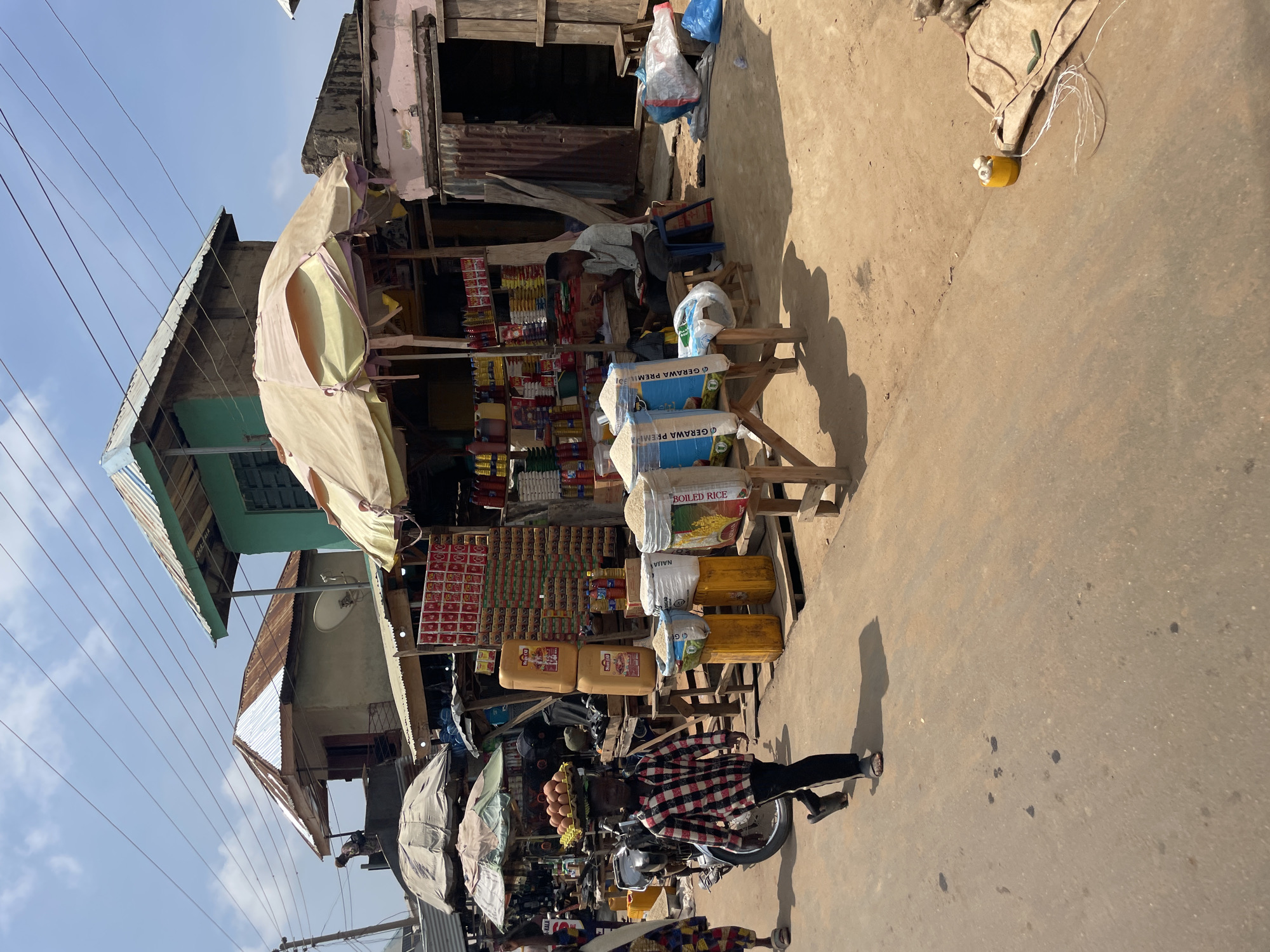 This image depicts a street-level view of a bustling market scene in Akwanga, Nigeria. The scene is dominated by a small shop or stall, characterized by its makeshift structure with a corrugated metal roof and weathered wooden supports. The stall is heavily laden with various goods, including sacks of rice, large yellow containers, and numerous smaller packaged items stacked on shelves. Some of the rice sacks have visible text, including "BOILED RICE" and "GERAWAY PREM".
In the foreground, a person wearing a red and black checkered shirt and black trousers is riding a motorcycle. The motorcycle is laden with a rack of eggs, suggesting this individual is a vendor or transporter of goods. Another person, partially obscured, is seated in the background near the stall, appearing to be resting. The ground is a dusty, unpaved surface, typical of many market areas in Nigeria.
Overhead, electrical wires crisscross the sky, and several buildings with similar corrugated metal roofs are visible in the background, indicating a densely populated urban or semi-urban environment. The lighting suggests it is daytime, with clear skies and sunlight casting distinct shadows on the ground. The overall impression is one of daily commerce and activity within a vibrant, albeit somewhat unrefined, marketplace. The setting is informal and practical, with goods displayed openly for sale.