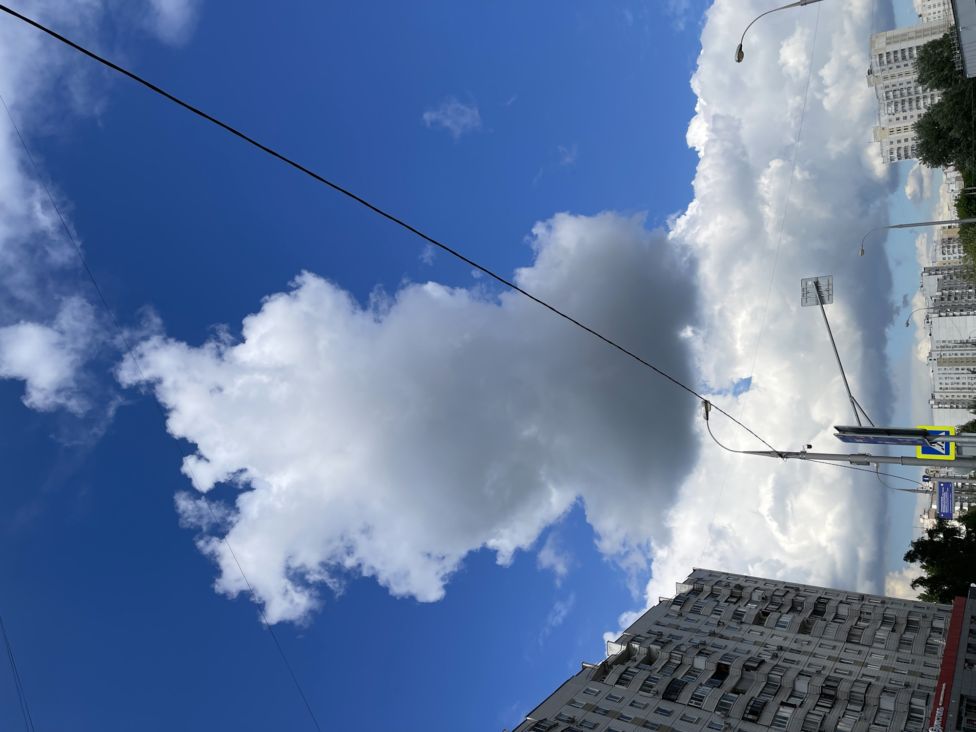 The image depicts a view looking upwards in a city, possibly Moscow, Russia. The sky is visible, displaying both clear blue areas and large cumulonimbus clouds. Some of the clouds appear dark gray, suggesting potential rain. Electrical wires stretch across the sky. The background shows multi-story apartment buildings, typical of urban residential areas. Streetlights and road signs are visible, further suggesting an urban environment. The buildings exhibit a repetitive pattern of windows and balconies.