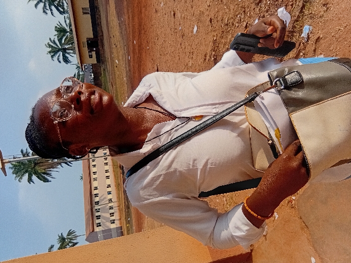 A woman with glasses and braided hair stands outside. She wears a white shirt and carries a two-toned purse. She holds a black smartphone. Palm trees are visible in the background, along with a tall, light-colored building with repeating windows. The ground appears to be reddish-brown soil. The lighting suggests a sunny day.