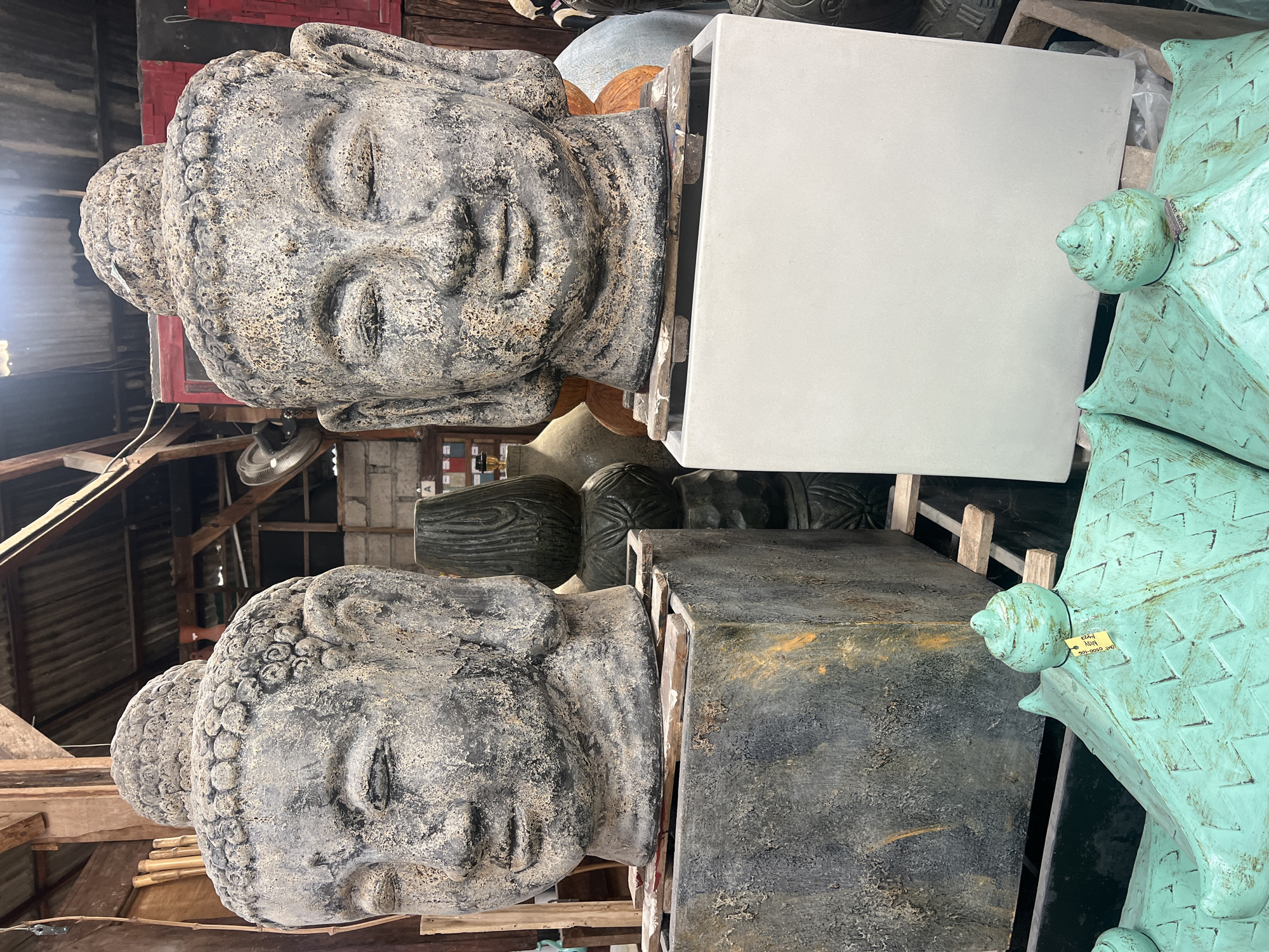 Two large, weathered Buddha heads made of stone or concrete are prominently displayed in an outdoor market stall in North Kuta, Indonesia. The Buddha heads are positioned on square pedestals, one dark gray and textured, the other a clean white. The head on the left has a slightly more weathered appearance with visible cracks and chips. Behind the Buddha heads, various other decorative items are visible, including stacked cylindrical vases with intricate carvings and what appear to be roof tiles with a green patina. The background consists of wooden beams and corrugated roofing, suggesting a covered but open-air market setting. A white tag is attached to the Buddha head on the right.