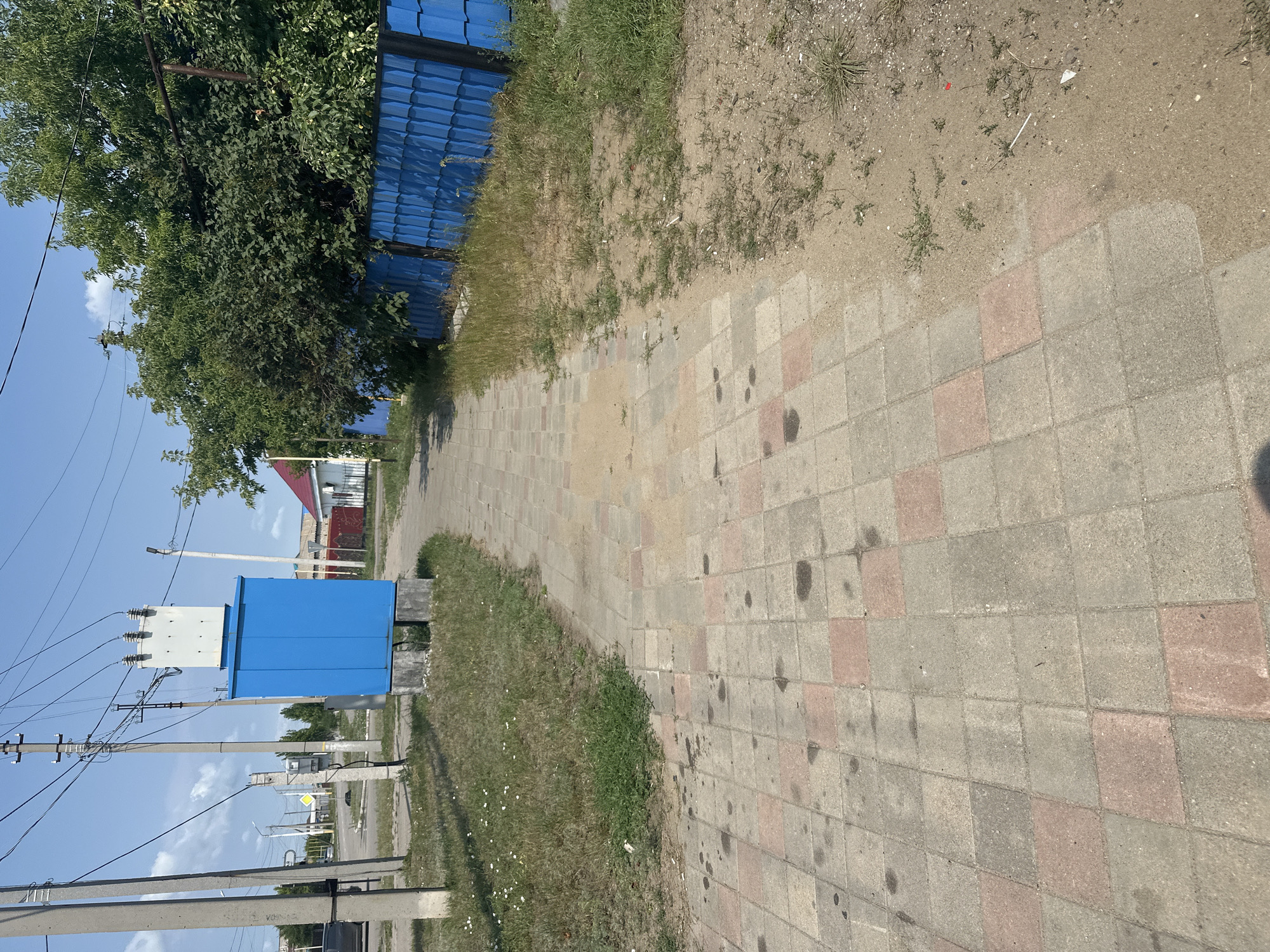 The image shows a paved sidewalk in Tobyl, Kazakhstan, leading towards a blue transformer box with a white electrical unit on top. The sidewalk is made of square concrete tiles in shades of gray and pink, with some dark stains. To the left of the sidewalk, there is a grassy area with some small white flowers and a concrete power pole with electrical wires. Behind the transformer box, there are some buildings and trees. To the right of the sidewalk, there is a blue metal fence and more trees. The sky is blue with some clouds. The scene appears to be a residential or suburban area. There are no people visible in the image.