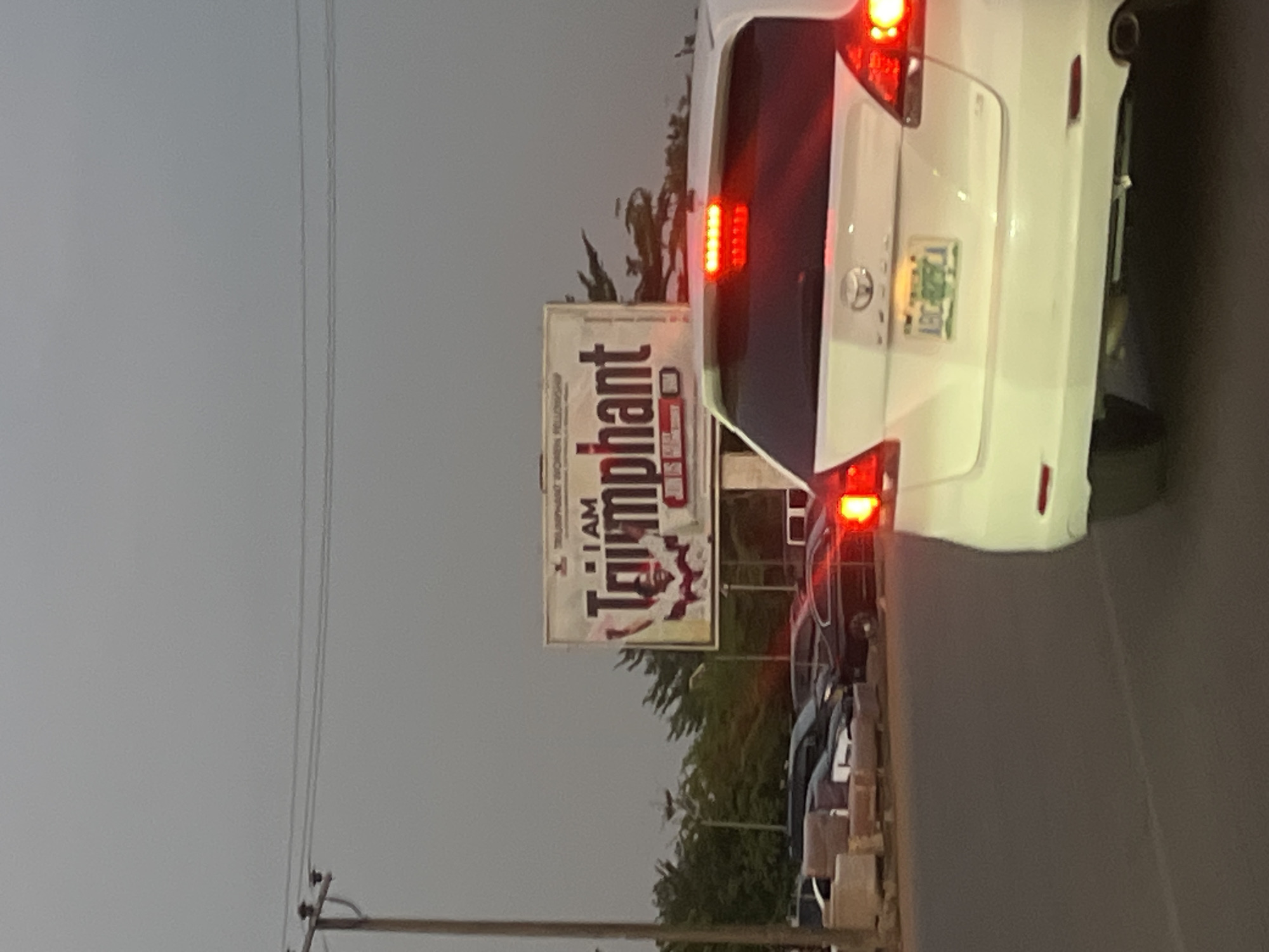The image captures a street scene in Galadimawa Village, Nigeria. A white SUV is positioned in the foreground, with its taillights illuminated. Adjacent to the vehicle, a billboard displays the text "I AM Triumphant," with the word "Triumphant" appearing larger and in a bolder font. The billboard seems to be an advertisement for a religious organization. Further down the street, a row of parked cars can be seen. The sky is overcast, suggesting that it might be late afternoon or early evening. A utility pole with power lines runs along the left side of the image.