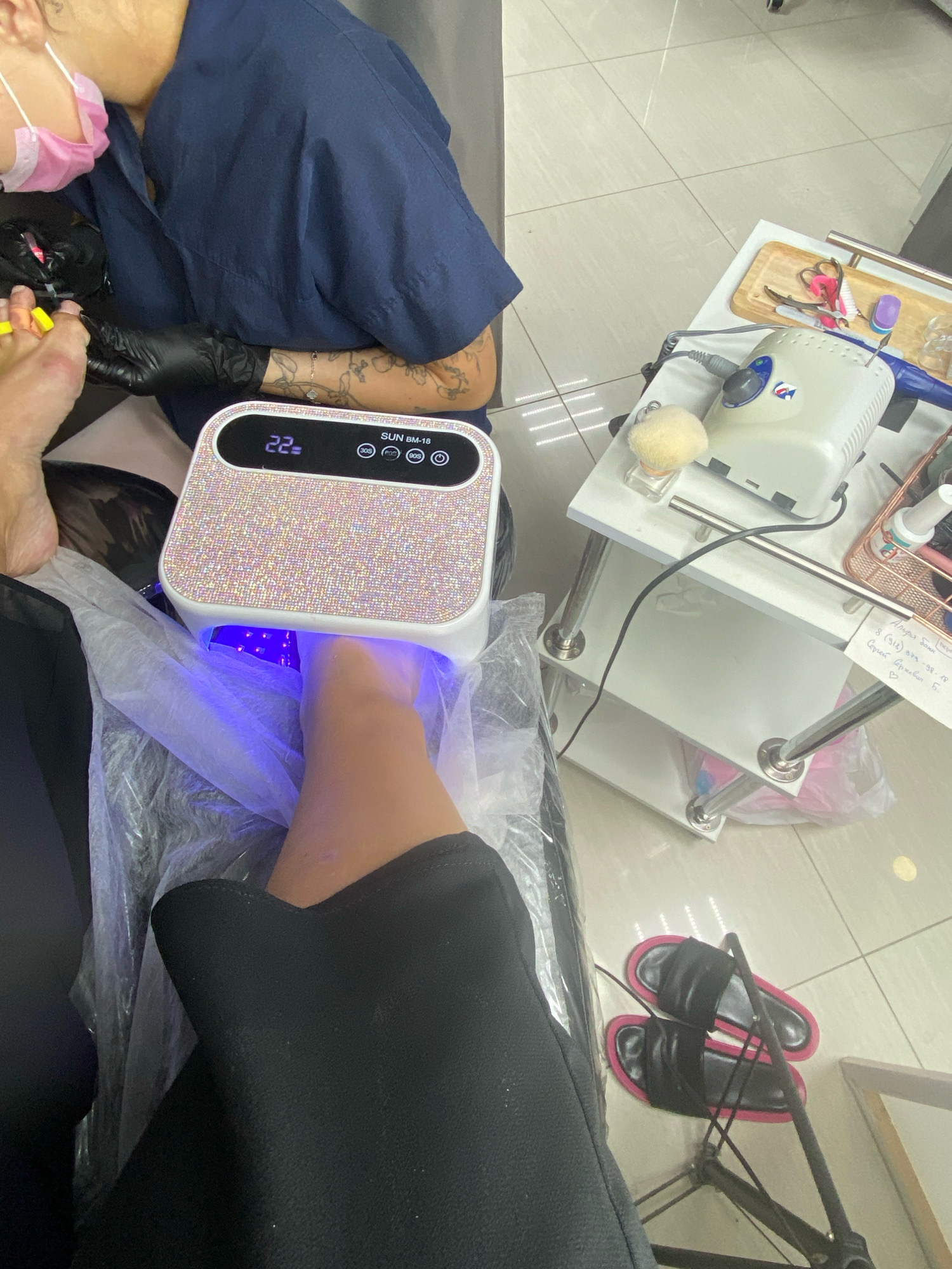 The image shows a close-up of a person's foot undergoing a nail treatment, with a technician working on it. The foot is placed inside a sparkly UV nail lamp, emitting a blue light. Yellow nail separators are visible on the toes. The technician, wearing a navy blue uniform and black gloves, is leaning over the foot, holding tools. Nearby, a white nail drill machine and other manicure tools are set up on a white trolley. On the floor, a pair of black and pink slides are visible. The scene appears to be in a nail salon, possibly located in Sochi, Russia, given the writing on a piece of paper attached to the trolley which contains Cyrillic script that appears to be Russian.