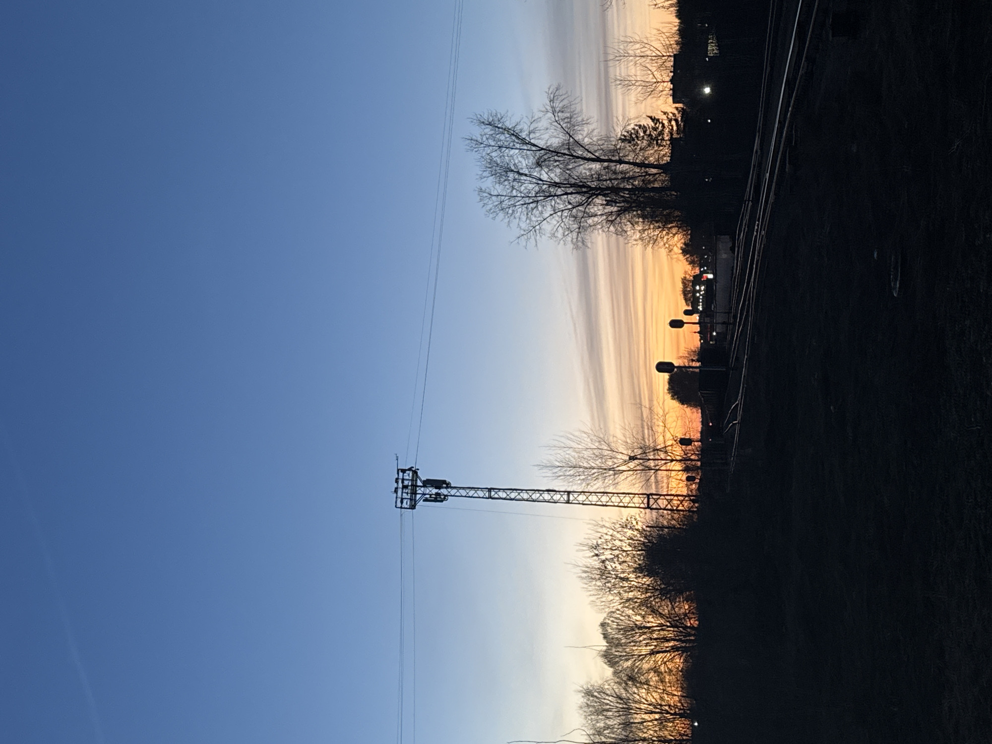 The image shows a landscape at dusk or dawn. The sky transitions from blue to orange, indicating the time of day. A utility tower stands prominently against the sky, with wires extending horizontally. Below the horizon, there are bare trees and vegetation, silhouetted against the fading light. A set of train tracks runs along the right side of the image. The location is Chaikovsky, Russia, based on available metadata and context. Details regarding people, specific actions, and interactions are not discernible from the image.