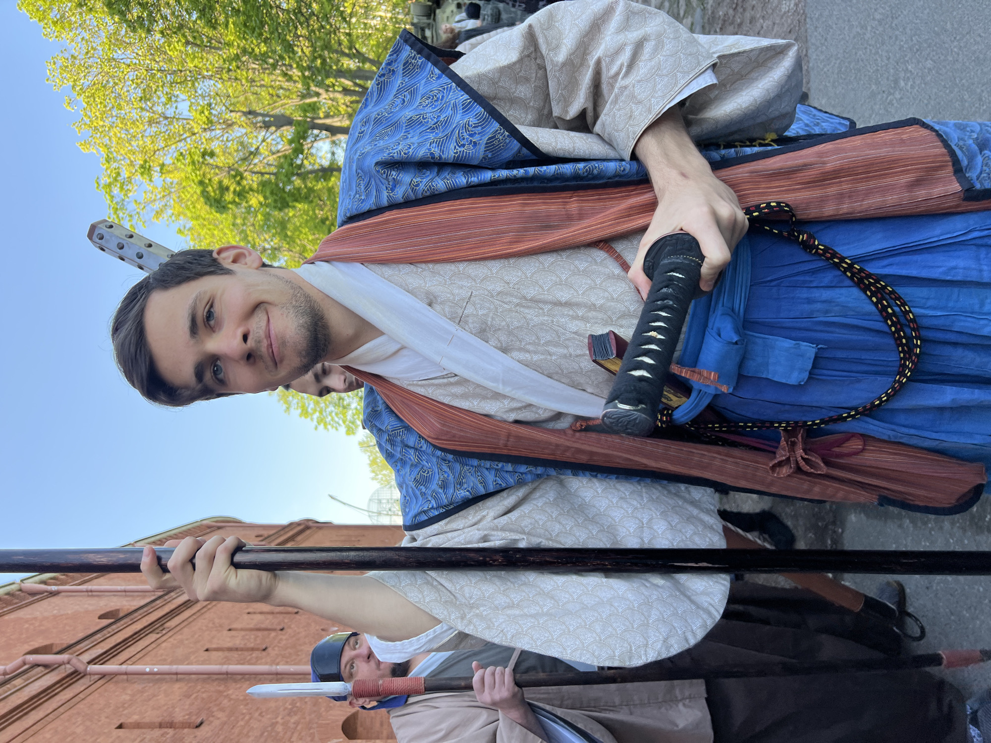 A sideways, medium shot shows a man in what appears to be a Japanese costume, holding a sword in his right hand. The man has short, dark hair and a light complexion, and he smiles slightly. His outfit includes a blue patterned outer layer over a cream-colored patterned kimono, with a brown sash. A white neck covering is visible. In the background, there are trees and a clear sky. Another person is partly visible behind him. Below, a group of people is holding long spears in front of a red brick building. The building appears to be the Saint Petersburg Fortress in Russia.