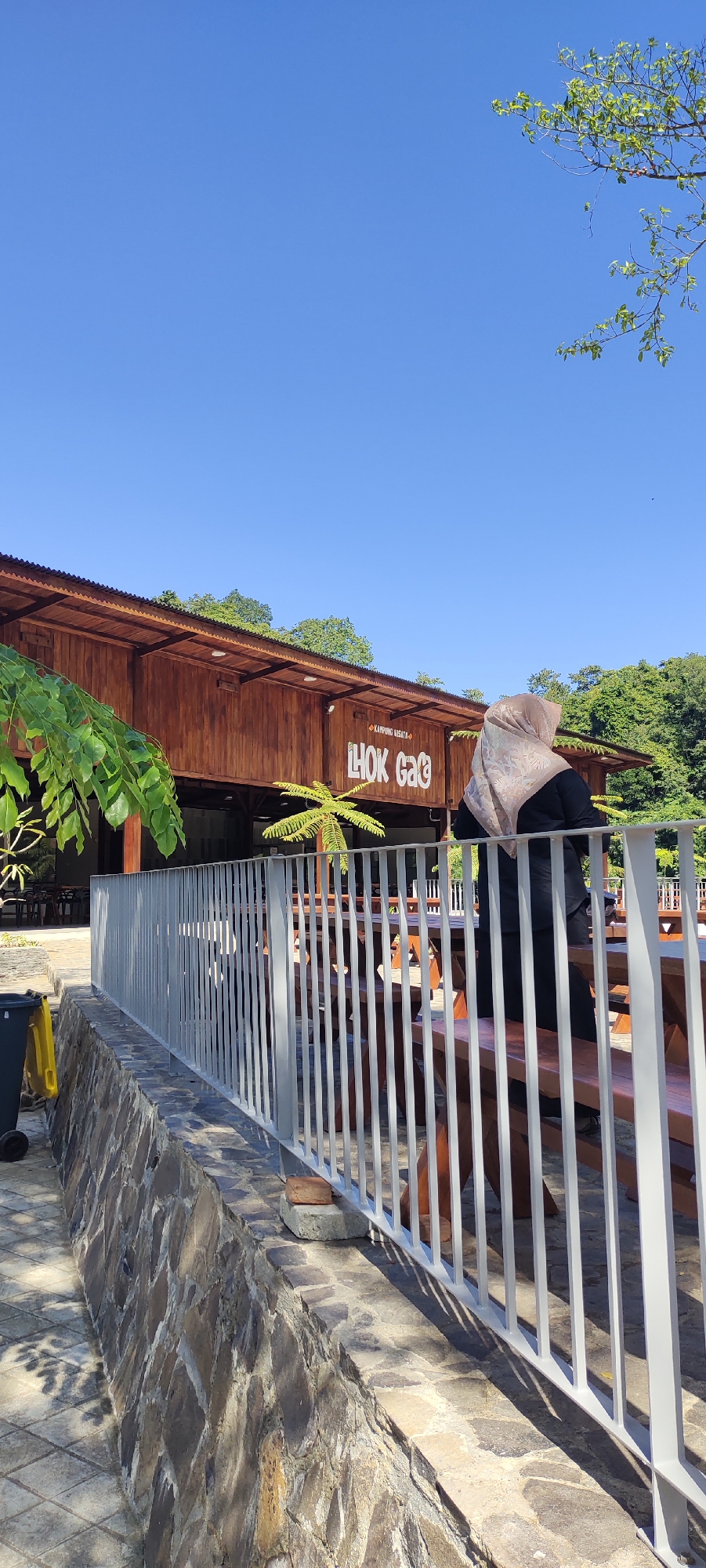 The image depicts the exterior of a wooden building with a corrugated metal roof, likely a cafe or restaurant, labeled "LHOK GAQ KAMPUNG WISATA" in large white lettering. The building is situated in a bright, sunny outdoor environment with a clear blue sky. In the foreground, a white metal railing runs along a low stone wall, separating the viewer from a seating area with wooden picnic tables and benches. A person wearing a beige hijab and a black outfit is visible from behind, standing near one of the tables. Lush green foliage frames the scene, with trees and branches visible against the sky and behind the building. To the left of the railing, a black trash can with a yellow lid is present. The ground is paved with light-colored square tiles. The overall impression is that of a scenic outdoor dining or relaxation spot.