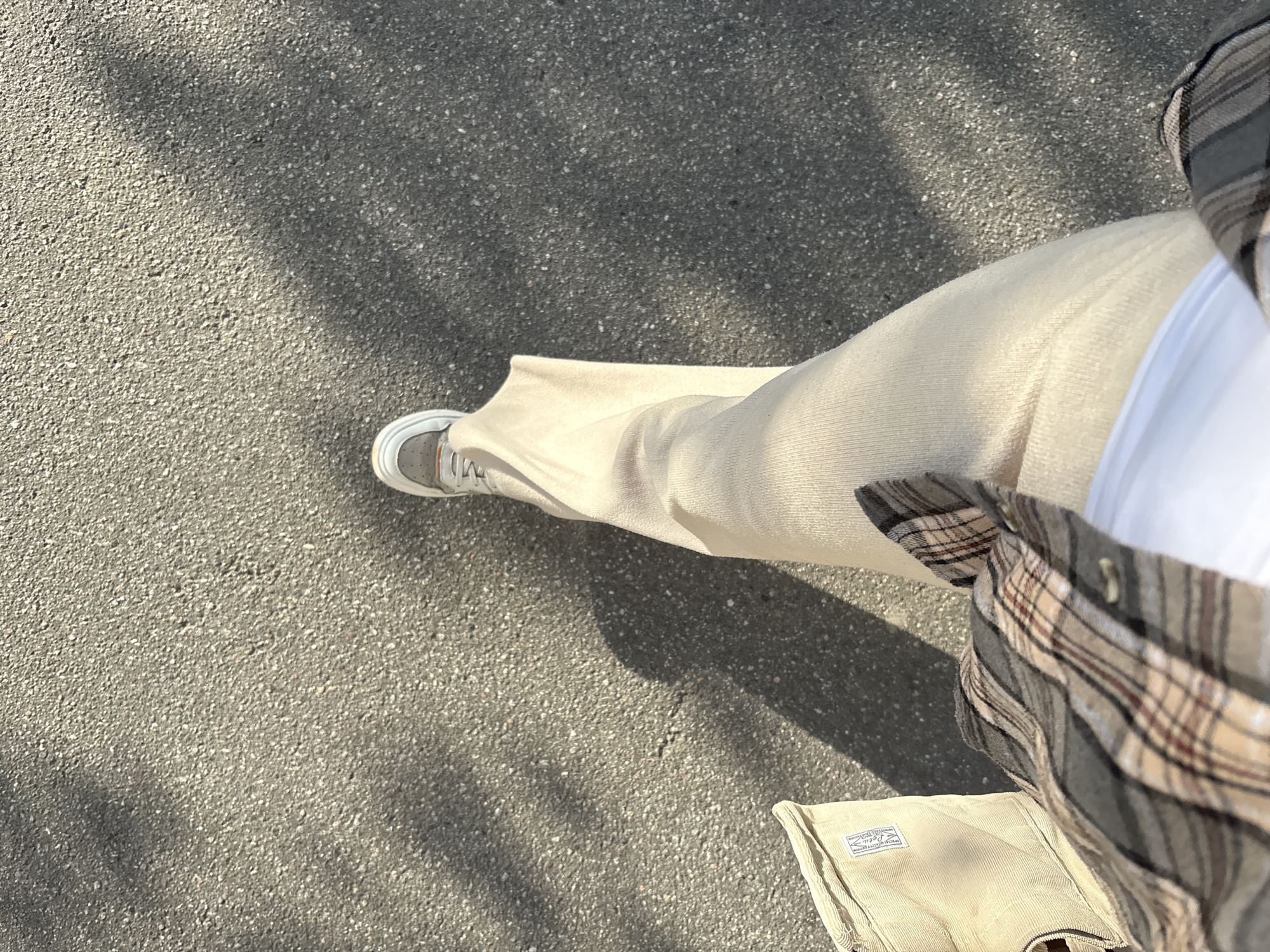 The image shows a person's legs and feet, as well as a portion of a plaid shirt and a corduroy bag. The person is wearing light-colored wide-leg pants and white sneakers with gray and orange accents. The plaid shirt is composed of gray, beige, and reddish-brown stripes. The corduroy bag is light beige and has a label with text on it. The ground is asphalt, with shadows of leaves cast across it, suggesting it is daytime with sunlight. The scene appears to be outdoors, possibly a street or sidewalk. The image is taken from a high-angle perspective, looking down at the person's legs. No specific activity is discernible other than the person appearing to be standing or walking. The overall impression is casual and relaxed.