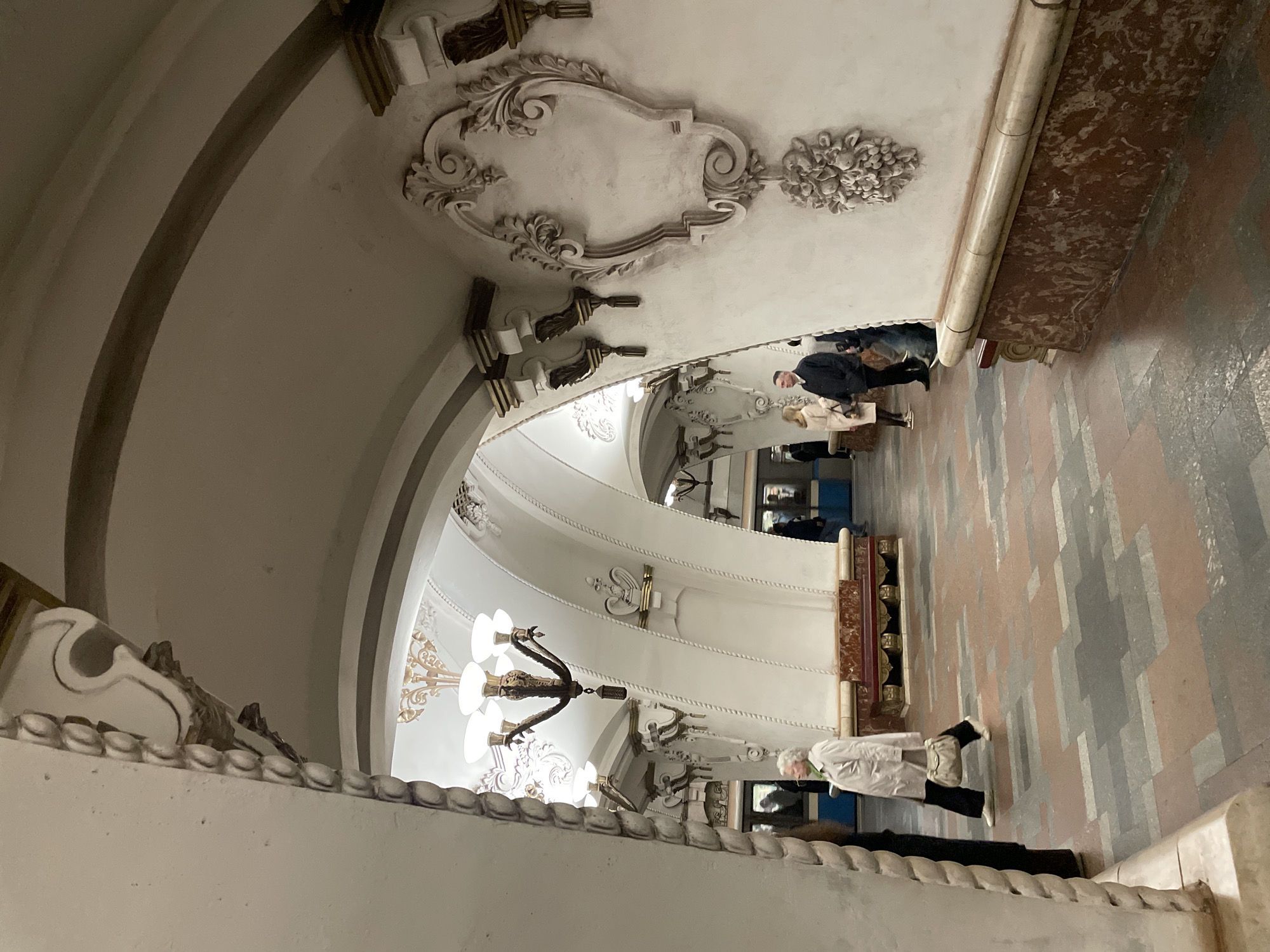 The image shows the interior of a Moscow Metro station, likely Kiyevskaya on the Arbatsko-Pokrovskaya line based on the ornate architectural details. The scene features an arched ceiling with decorative moldings and plasterwork, including floral and frame-like embellishments. A multi-tiered chandelier hangs from the ceiling. People are present, including individuals walking along the tiled floor. The flooring has a patterned design. Some people are waiting at a wall that has some type of sculpture design at the top. The color palette is primarily muted shades of white, gray, and brown.