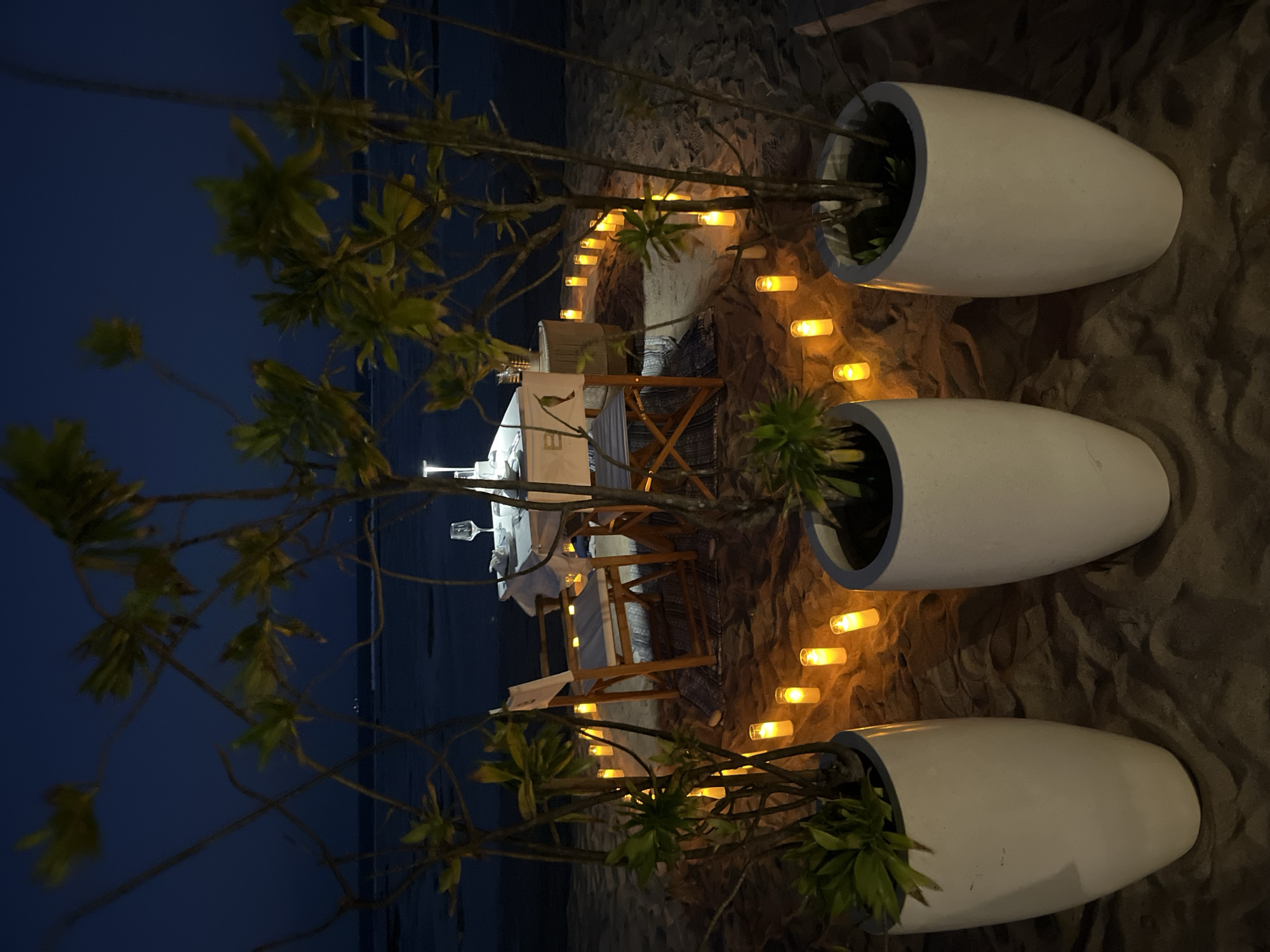 A romantic, candlelit dinner for two is set up on a sandy beach at dusk in Kuta Selatan, Indonesia. The scene features a round table laden with white linens, a wine glass, and other tableware, illuminated by a central lamp and a perimeter of glowing yellow candles. Two chairs, one with a white canvas backing displaying the letter "B", are positioned at the table. The setting is further enhanced by three large white planters, each containing a small, leafy green plant, arranged in the foreground. The calm ocean and the darkening sky form the backdrop. The image captures a tranquil and intimate dining experience by the sea.