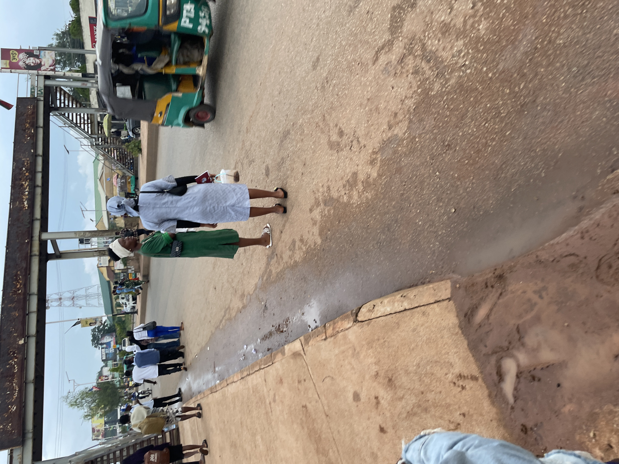 This is a street-level photograph taken in Jos, Nigeria. The image captures a moment on a dusty road under an overcast sky, possibly during or after a rain shower, as evidenced by the puddles and dampness on the asphalt and the nearby concrete walkway.
In the foreground, a person's legs, clad in light blue denim jeans, are visible from the knee down, suggesting they are either standing on the walkway or in the process of walking. The walkway itself is made of reddish-brown concrete and appears somewhat worn. Adjacent to the walkway is a roadside drainage channel, which is partially filled with muddy water and debris.
Moving into the midground, two women are the most prominent figures. The woman on the left wears a vibrant green dress and a white headwrap. She is facing away from the camera, looking towards the street. The woman on the right is dressed in a light blue, long-sleeved dress and also wears a white headwrap. She is also facing away from the camera. She carries a white bucket in her right hand and what appears to be a notebook or small book in her left hand.
Further into the background, several other individuals are present. On the left side of the street, a group of people, mostly men, are gathered. One man in the foreground is wearing a white shirt and patterned shorts and carries a backpack. Another individual is wearing a white shirt and dark trousers. A man in a blue shirt and black trousers is also visible. There's a sense of them either waiting or preparing to cross the street.
To the right of the road, a distinctive green and yellow auto-rickshaw, also known as a Keke Napep, is in motion, its front wheel blurred by the movement. The vehicle has the registration number "PTA 9450" partially visible on its side. The auto-rickshaw appears to be carrying passengers.
The background reveals elements of the urban environment, including a pedestrian bridge spanning across the road, characterized by its metal structure and concrete steps. In the distance, there are buildings, utility poles, and telecommunication towers, typical of a city landscape. Advertisements are visible on some structures, indicating commercial activity. The overall impression is a busy street scene in a Nigerian city.