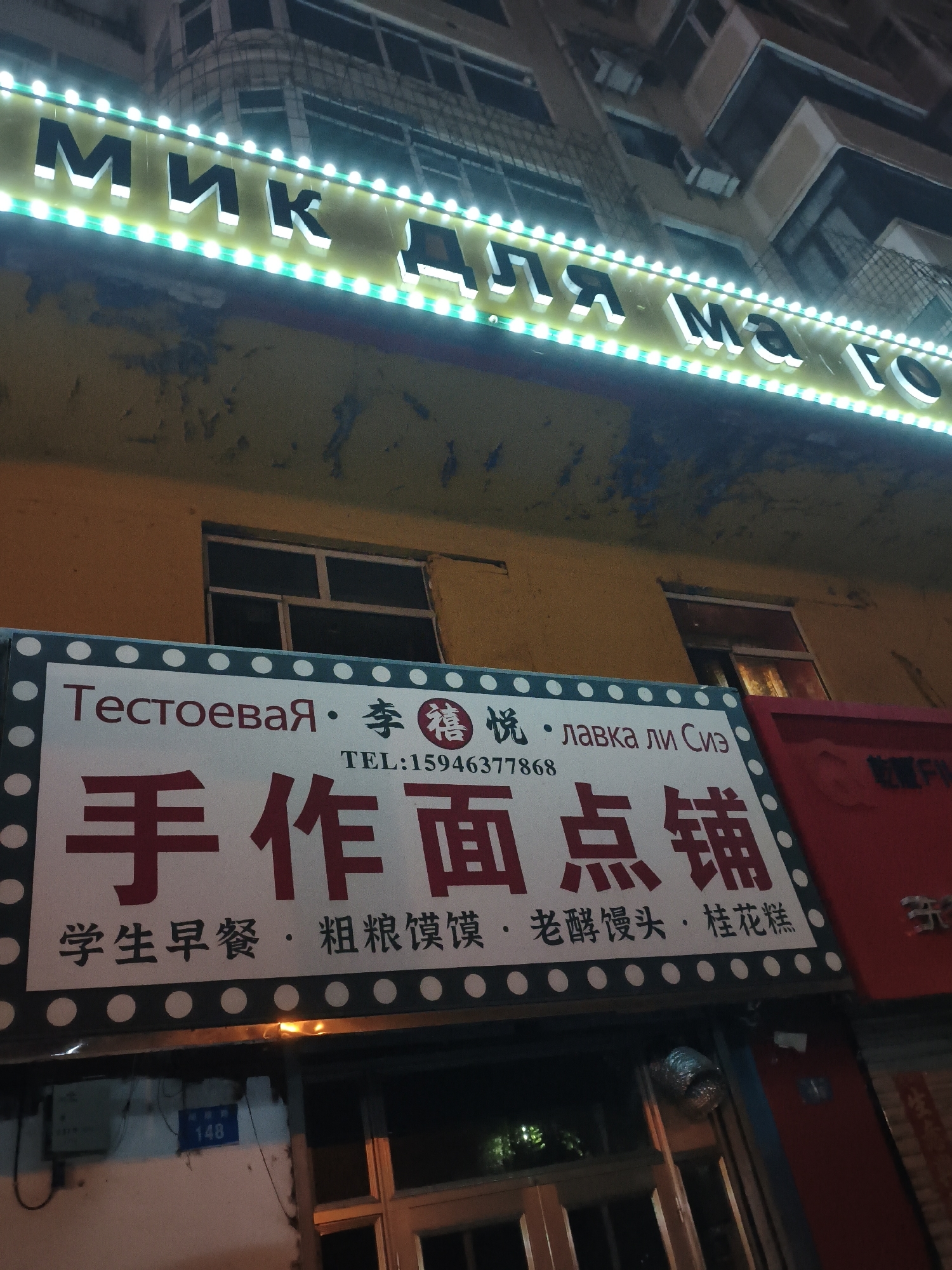 The image depicts a night scene in Xi'an, China, focusing on the facade of a building with illuminated signage.
Dominating the upper portion of the image is a bright neon sign with white, three-dimensional letters outlined in green. The sign appears to be in Cyrillic script, spelling out "МИК ДЛЯ МаГо", which translates roughly to "MIC FOR MAGO". The letters are large and stand out against the dark background.
Below this, a rectangular sign with a white background and a dark blue border is prominently displayed. This sign features text in both Russian and Chinese. The Russian text reads "Тестовая · 李 悦 · лавка Ли Сиз" and "TEL:15946377868". The Chinese characters on this sign are large and red, reading "手作面点铺", which translates to "Handmade Noodle and Dim Sum Shop". Below this, in smaller red Chinese characters, are the words "学生早餐 · 粗粮馒头 · 老酵馒头 · 桂花糕", meaning "Student Breakfast · Coarse Grain Mantou · Old Yeast Mantou · Osmanthus Cake".
The building itself has a yellow-orange facade, showing signs of age and peeling paint. There are dark-paned windows visible on the upper floors and at street level. To the right of the main sign, a red sign with some indecipherable text is partially visible.
At the bottom left, a grey electrical box is mounted on the wall, with a blue sign below it showing the number "148". The overall environment suggests an urban setting at night, likely a commercial area. There are no people visible in the frame. The lighting is dominated by the bright neon signs, creating a contrast with the darker building and surroundings.