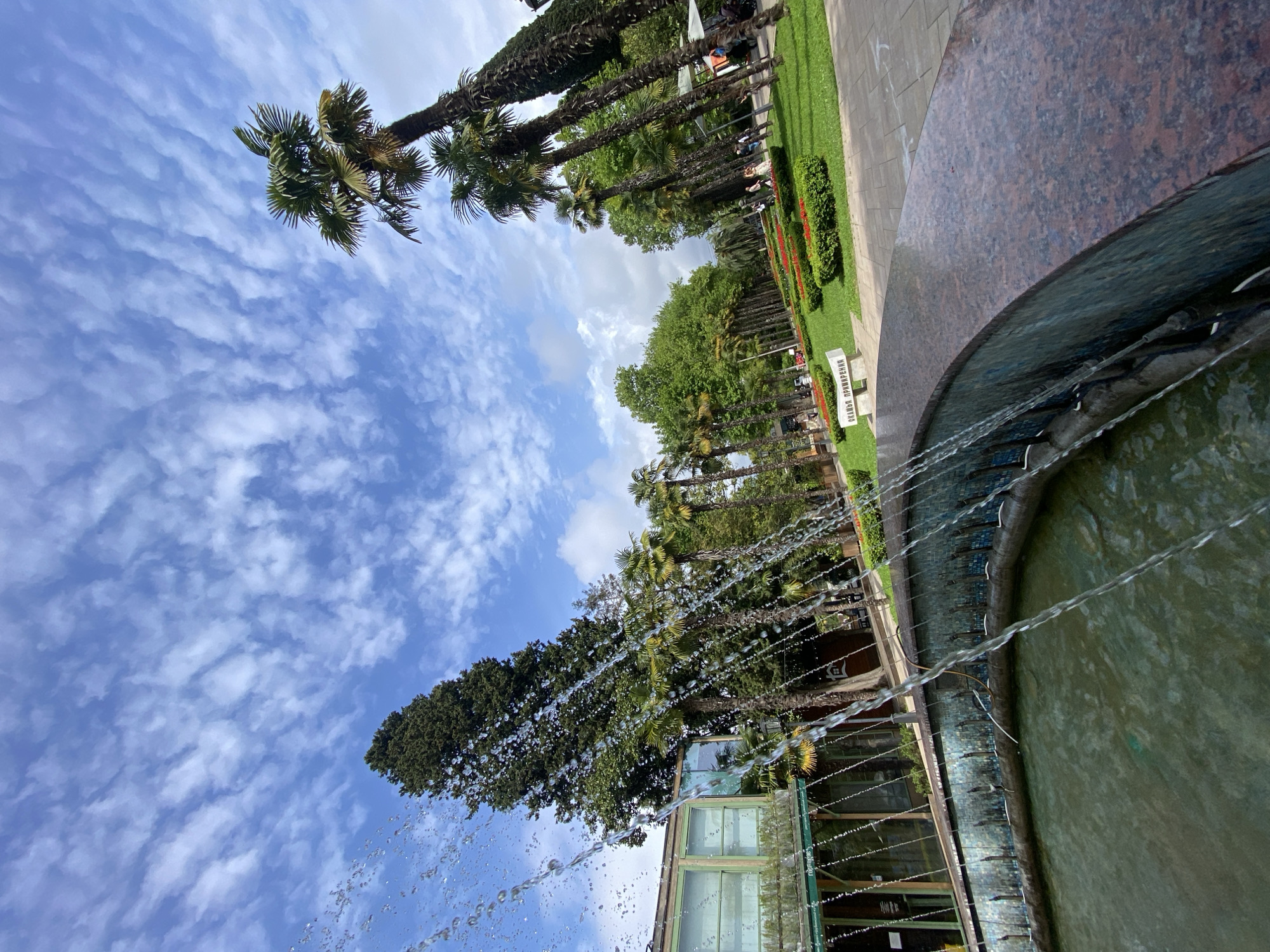 The image depicts a fountain in the city of Sochi, Russia. The fountain's edge is made of granite and blue-green tiles. Water is cascading from the edge of the fountain into its basin. Beyond the fountain, palm trees and other greenery line a walkway. A white bench is visible along the walkway. A building with green window frames is seen partially obscured by trees. The sky is blue with textured white clouds.
