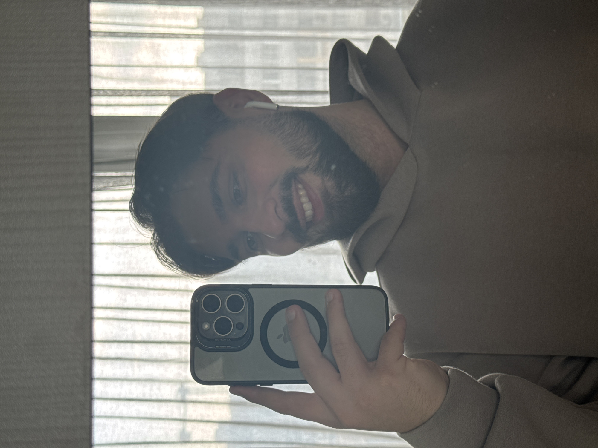 A man with dark hair and a beard smiles while taking a selfie in front of a window with closed blinds. He is wearing an off-white hoodie. He is holding a smartphone with a gray case and a circular magnetic attachment. The background appears to be an interior space near a window. There is no discernible information in the image to indicate a specific location in Varna, Bulgaria.