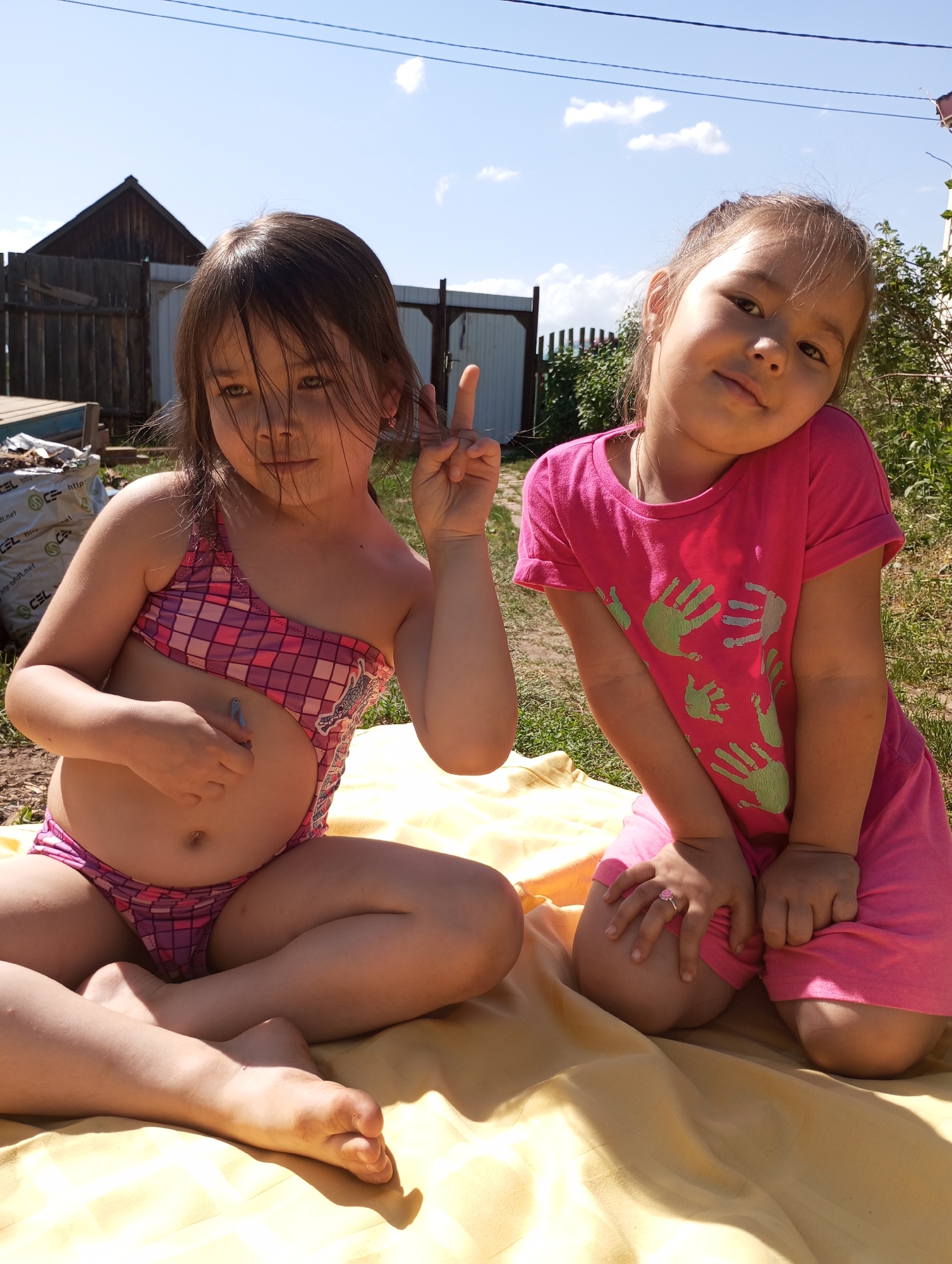 Two young girls are sitting on a yellow blanket outdoors on a sunny day. The girl on the left is wearing a pink and purple checkered swimsuit and holding up two fingers in a peace sign. The girl on the right is wearing a pink t-shirt with green handprints and pink shorts; she is leaning forward with her hands on her knees. Behind them, there's a wooden house, a white fence, and some greenery. The location is likely in or near Буренка, Russia. There are electrical lines visible in the sky.
