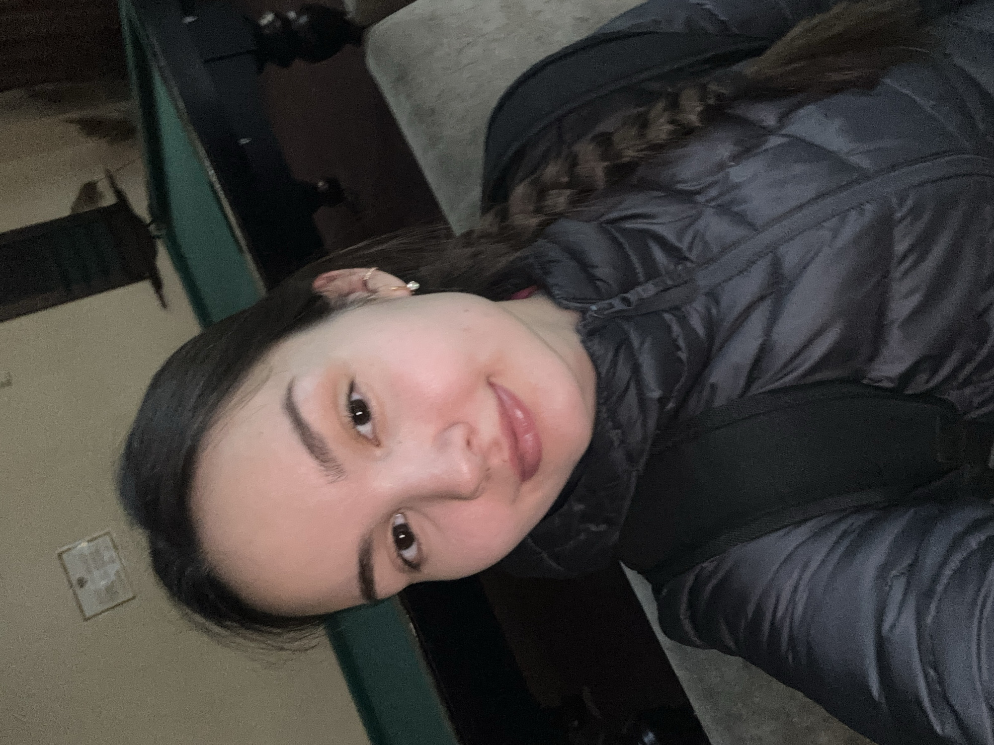 A young woman is pictured indoors. She has fair skin, dark hair, and brown eyes. She is wearing a dark puffer jacket. She is positioned with her head toward the left side of the frame. The background includes a green-painted object and an electrical outlet. The location is Orenburg, Russia.