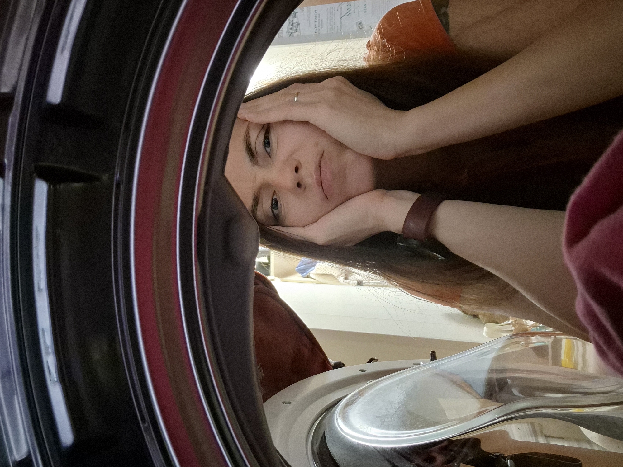 An indoor, eye-level, slightly upward-tilted shot captures a woman looking directly into the lens from the perspective of a front-loading washing machine's drum. The woman, appearing to be in her 20s or 30s, has long brown hair and blue eyes. She wears an orange/rust-colored top. Her hands cup her face; one hand rests on her forehead, and the other supports her chin, conveying a pensive, slightly weary, or bored expression. A plain ring is visible on her left ring finger, and a dark brown wristband is on her right wrist.
The foreground on the left features the dark, curved interior components of the washing machine, likely the door gasket and inner rim. On the bottom right, parts of the washing machine's white frame and reflective silver elements are visible, possibly from the detergent drawer or control panel. The background behind the woman shows a light-colored wall, possibly adorned with faintly discernible, reversed text or a patterned wallpaper.
The scene suggests a domestic setting, likely a laundry area within a home in Chaikovsky, Russia. The woman's pose indicates she is actively engaged with or observing the washing machine, possibly during a laundry cycle. The lighting suggests daytime indoors. No explicit text is clearly legible.