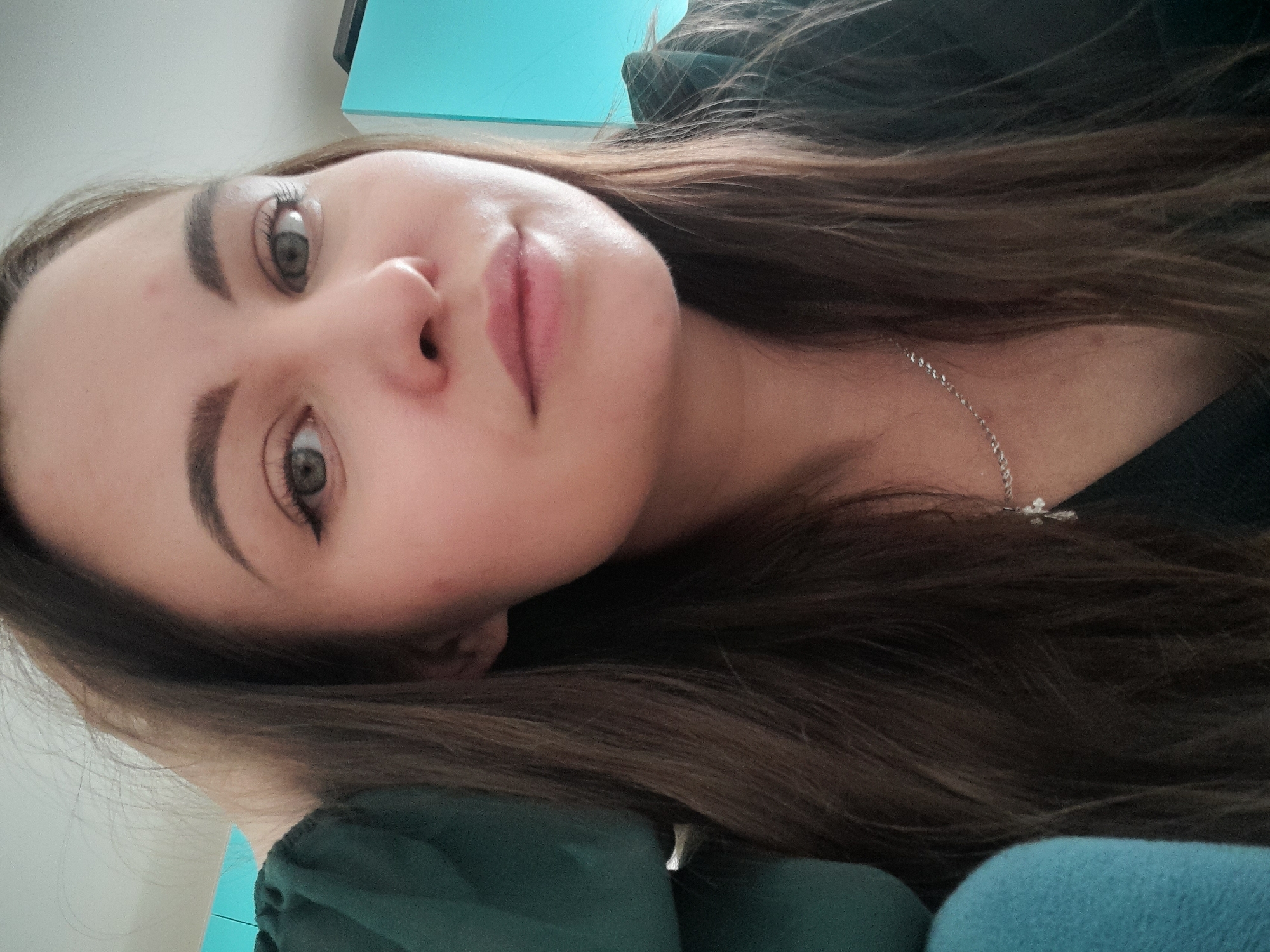 A close-up portrait of a woman with fair skin, dark eyebrows, and light green eyes. Her long, brown hair is slightly disheveled and falls around her face and shoulders. She is wearing a silver necklace with a pendant. The background includes a teal surface, possibly a cabinet or wall, and dark green fabric. The scene lacks information that would enable precise location.