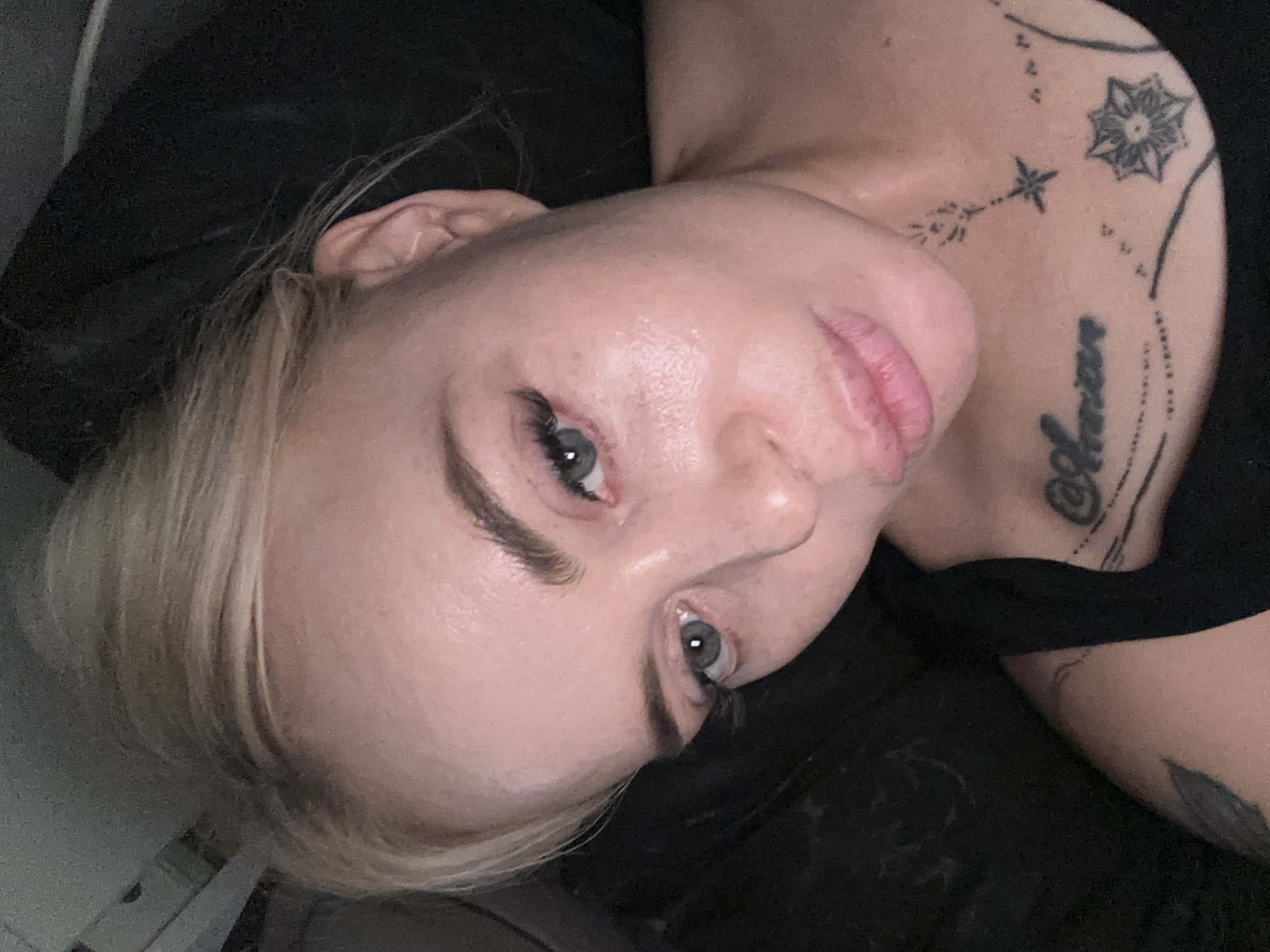 A close-up, over-the-shoulder selfie of a blonde woman is shown. She has light skin, blonde hair pulled back, and grey eyes with long false eyelashes. Her lips are slightly parted. She is wearing a black tank top. A tattoo of the word "Amiara" is visible on her chest, as well as other smaller star-shaped tattoos and designs on her neck and collarbone. The background is dark and out of focus, suggesting an indoor environment. The lighting is dim. The location is not specified.