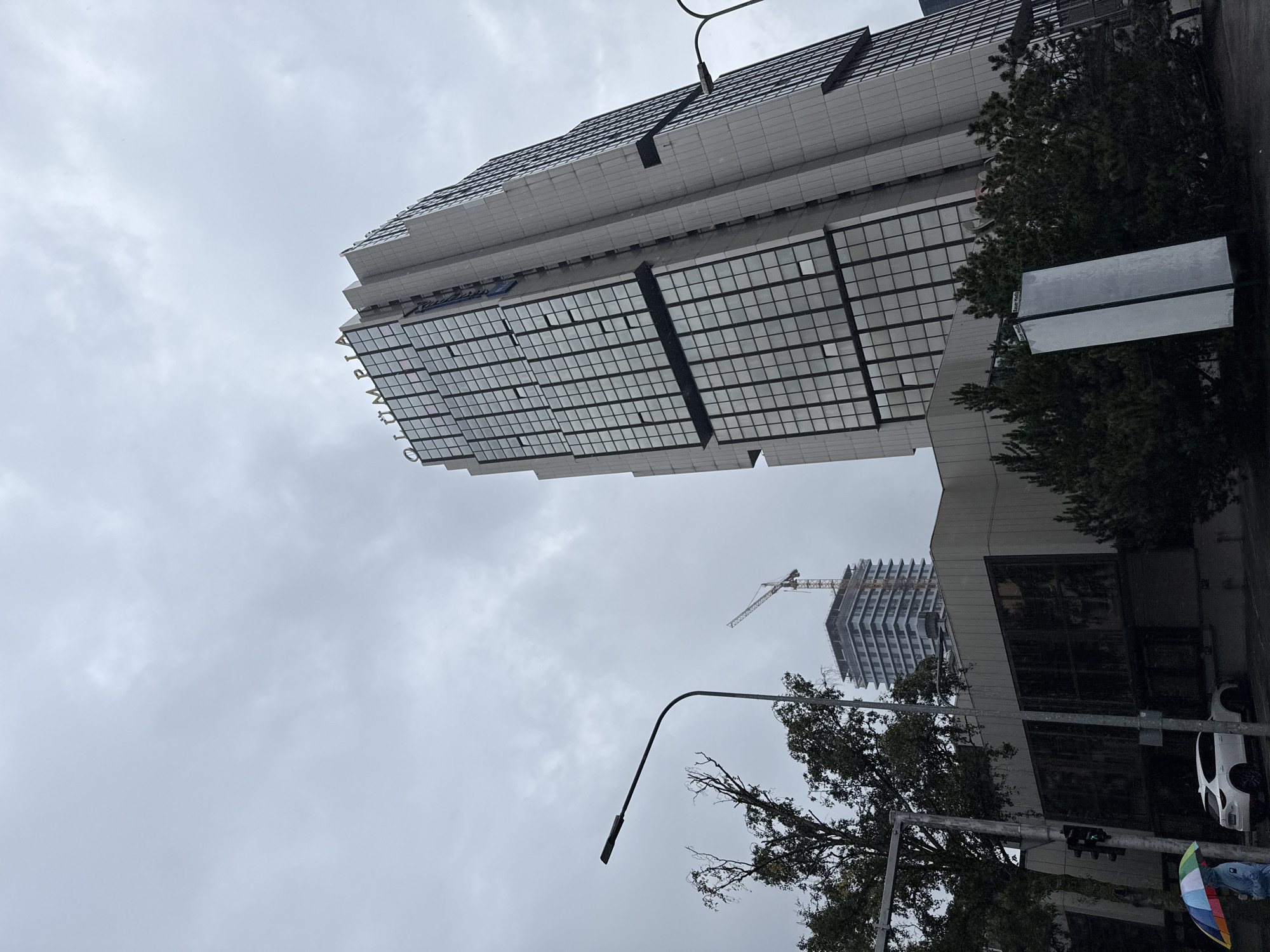 The image depicts a street view in Tallinn, Estonia, under a cloudy sky. In the foreground, a person holding a colorful umbrella walks past a white car. A traffic light is visible, showing a green signal. To the left, a tree with sparse branches is present. Further into the background, a tall, modern building with a glass facade and the word "OLÜMPIA" written in golden letters is prominently featured. To the left of the Olympia building, another taller building under construction is visible, with a crane on top. The overall atmosphere suggests an overcast day, possibly with light rain.