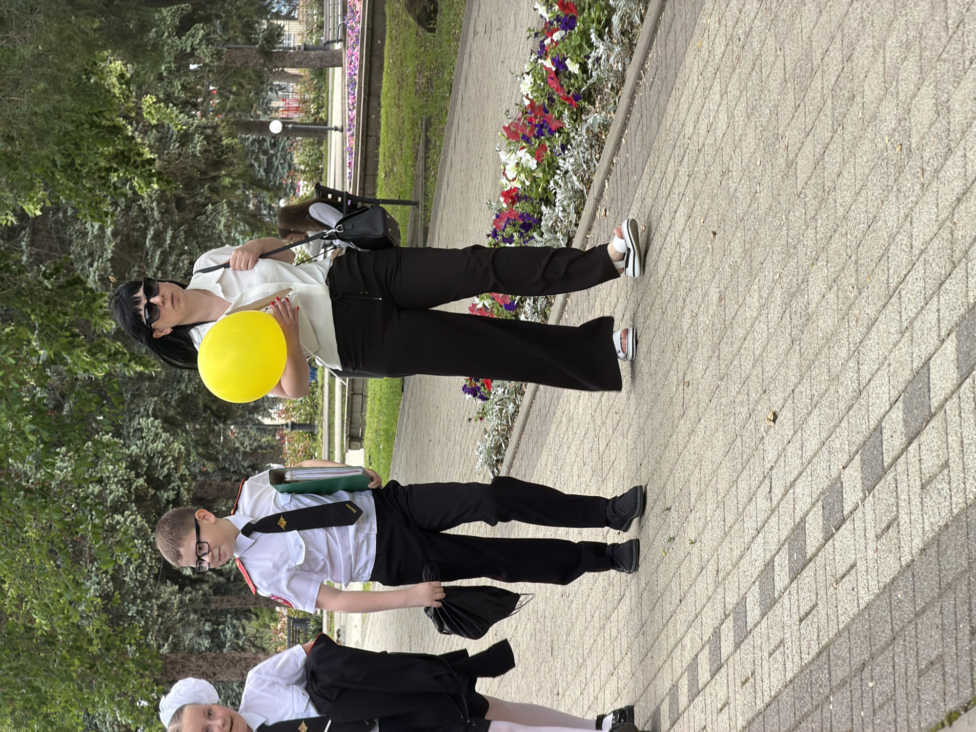 A woman with black hair, sunglasses, a white shirt, and black pants is holding a yellow balloon. She is standing on a brick walkway alongside a flower bed with red, white, and purple flowers. A boy wearing glasses, a white shirt, a black tie, and black pants stands next to her, holding green folders and a black cloth bag. A girl in a white bonnet, white shirt, and black skirt is partially visible near the bottom of the image. The background includes trees and a green grassy area. The image appears to have been taken in a park-like setting. There is no specific information about Leningradskaya, Russia.