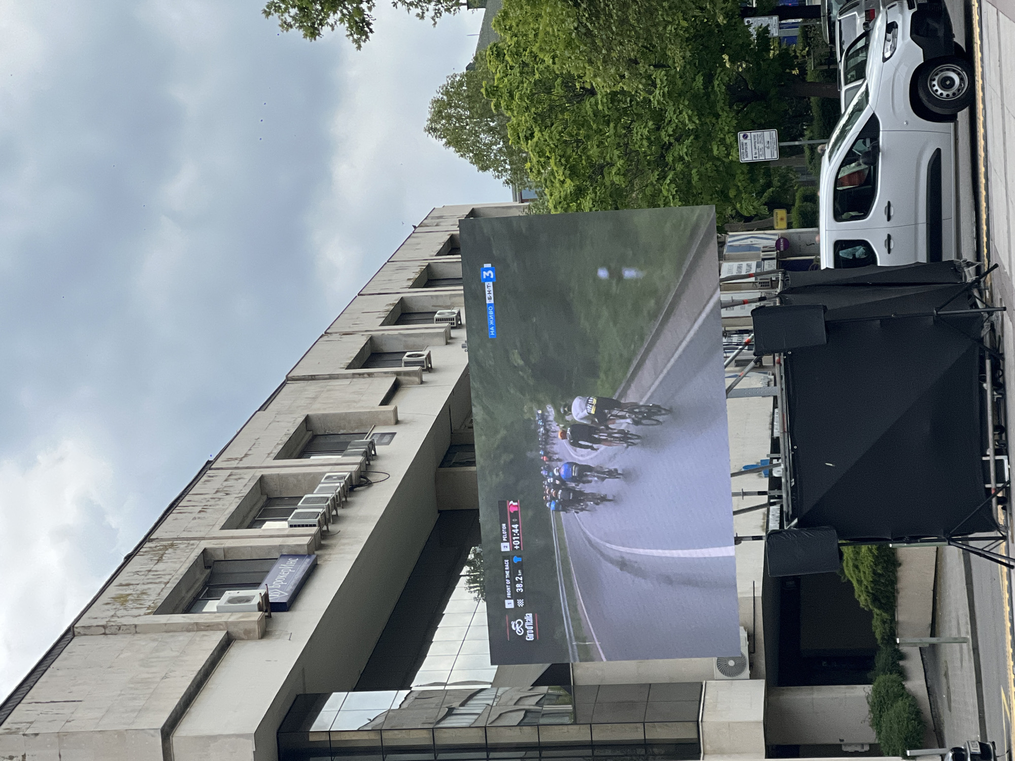 The image shows a large outdoor television screen displaying a live broadcast of a cycling race, specifically the Giro d'Italia, in Burgas, Bulgaria. The screen is positioned in front of a modern building with several windows and air conditioning units attached to its facade. A van is parked to the side of the screen. The race is being broadcast live on BNT 3, the national television channel of Bulgaria. The screen displays data showing the speed of the lead rider (38.2 km/h), the gap between the lead rider and the Peloton (+01:44), and the "ON AIR" designation on the screen. The logo of a company called "Брокеринс" is visible on the building. The weather on screen appears wet.