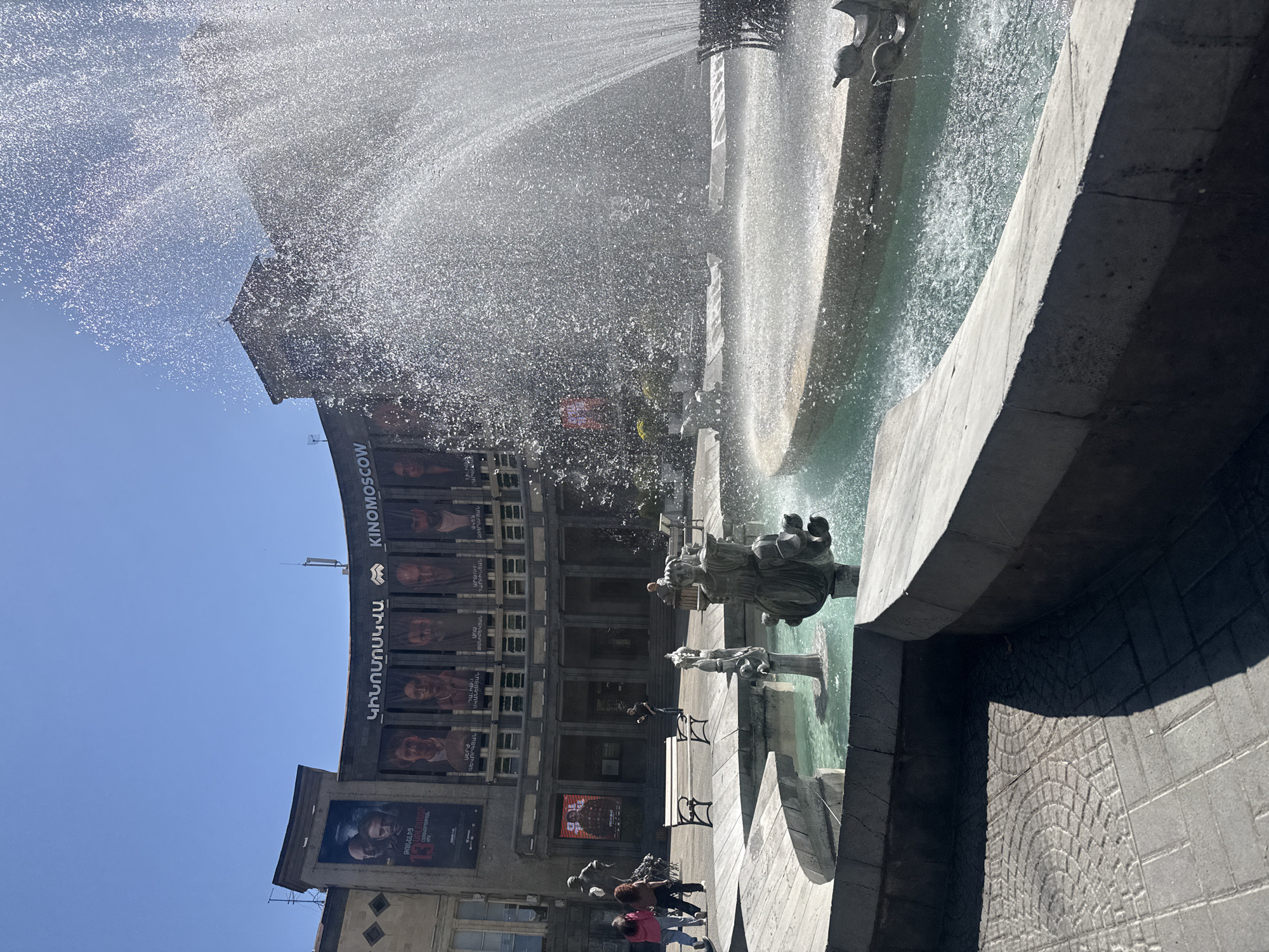 The image captures a daytime scene at Yerevan's Charles Aznavour Square in Armenia, featuring a prominent fountain with multiple tiers. Water sprays from the fountain, partially obscuring the view of the Kino Moscow cinema in the background. The cinema's facade is adorned with posters and the "KINOMOSCOW" sign written in both English and Armenian. Benches are placed near the fountain, and several people are present in the square, including a group walking near the base of the fountain. The sky is clear and blue. The fountain's structure and the surrounding pavement are made of stone or concrete.