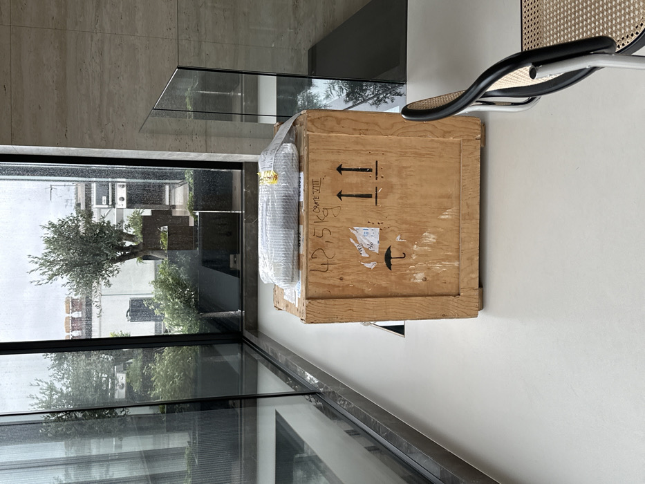 The scene depicts an interior space with a large, light-brown wooden shipping crate situated centrally on a smooth, light-colored floor. The crate is marked with "42.5 kg", "CRATE VIII", and international shipping symbols including two upward arrows for "this way up" and an umbrella for "keep dry", along with various white labels. Resting atop the crate is a rectangular item wrapped in clear bubble wrap and secured with tape, displaying a small golden label. To the right, the back and part of the seat of a modern chair with a black frame and woven cane material are visible.
The interior features large floor-to-ceiling glass windows, wet with rain, that form a corner and look out onto an exterior rooftop terrace. The terrace contains multiple dark rectangular planters holding mature olive trees and various shrubs. In the background, beyond the terrace, white buildings with visible chimney pots rise under an overcast sky, characteristic of architecture within the city of London, United Kingdom. No individuals are present in the scene, and no discernible actions or interactions are occurring.