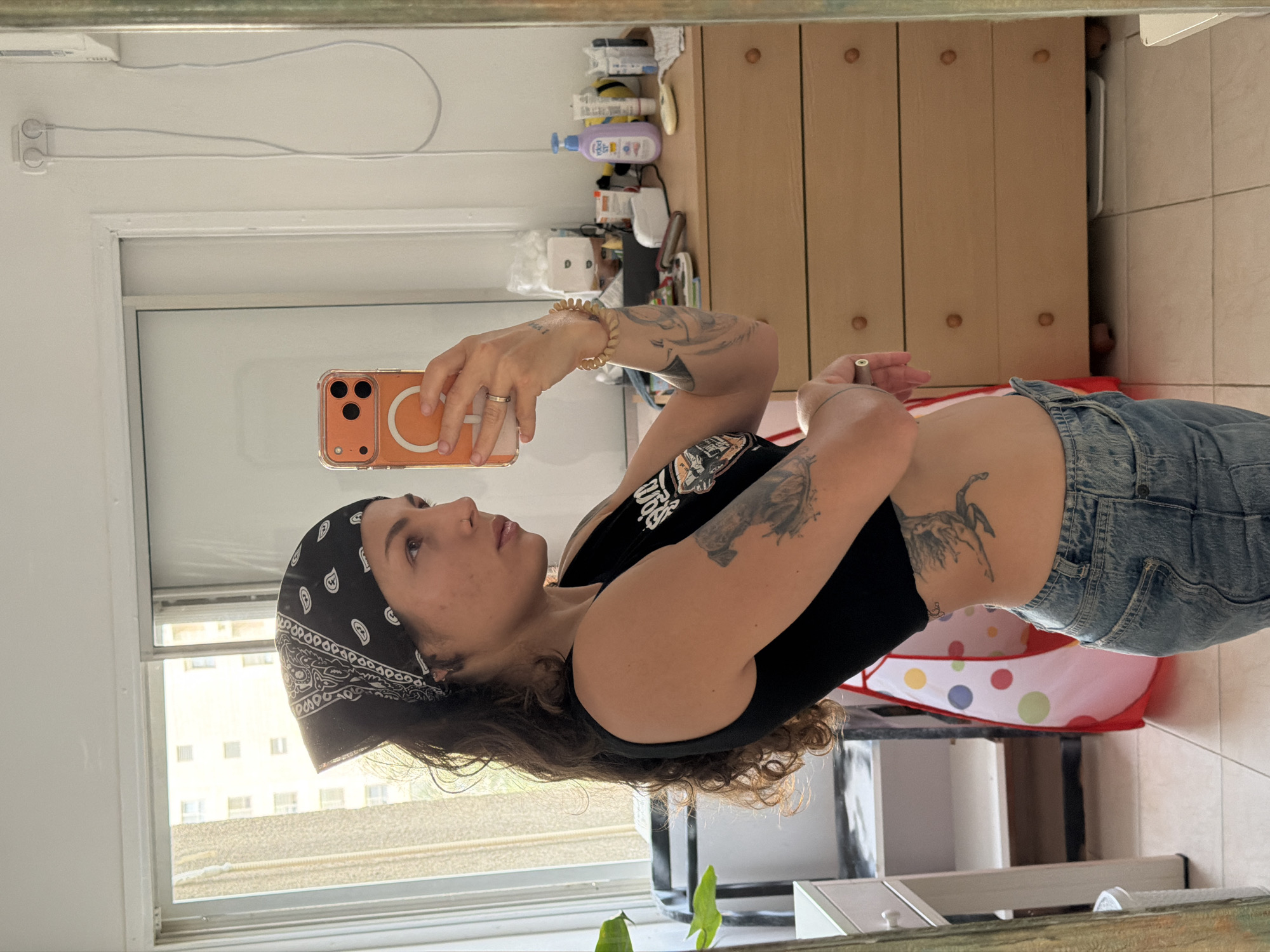A woman takes a photo of herself in a mirror. She wears a black tank top, denim jeans, and a patterned bandana on her head. Her arms are adorned with tattoos. She holds an orange phone in her left hand, taking the picture. The reflection shows a room with a window offering a view of a building complex. Inside the room is a light-colored wooden dresser and a colorful polka-dot playpen. Based on visual information only, I cannot determine if the photo was taken in Kiryat Bialik, Occupied Palestine.