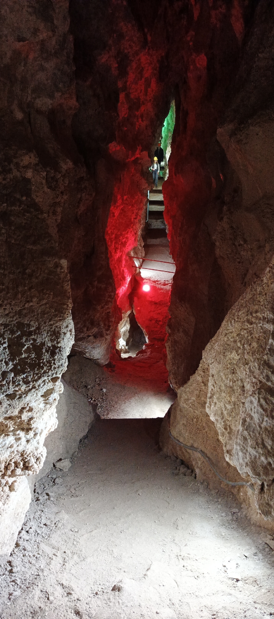 This vertical image depicts the interior of a dark, narrow cave passage, featuring rugged rock formations and an illuminated pathway. The foreground shows a dusty, sandy path with subtle white light revealing the ground texture and small stones.
Moving deeper into the cave, the passage narrows and appears to ascend, becoming dramatically lit by intense red light. This red illumination bathes the rough, irregular rock walls and parts of the path, creating a striking contrast with the surrounding darkness. A handrail, likely made of metal, is visible on the right side of this ascending section, offering guidance. Further along, a small, bright white light source is visible on the ground within the red-lit area.
In the distance, at the top of the illuminated ascent, two individuals are visible, appearing to climb steps or a steep ramp. One person is clearly seen wearing a yellow hard hat and a light-colored top, while the second individual is partially obscured behind them. Beyond these figures, a small opening or section of the cave is distinctly lit with a vibrant green light, providing a final color accent to the subterranean scene.
The constructed path, safety railings, and the use of contrasting colored artificial lighting suggest this is a developed tourist attraction or show cave. The activity is clearly cave exploration or a guided tour. No visible text is present in the image.