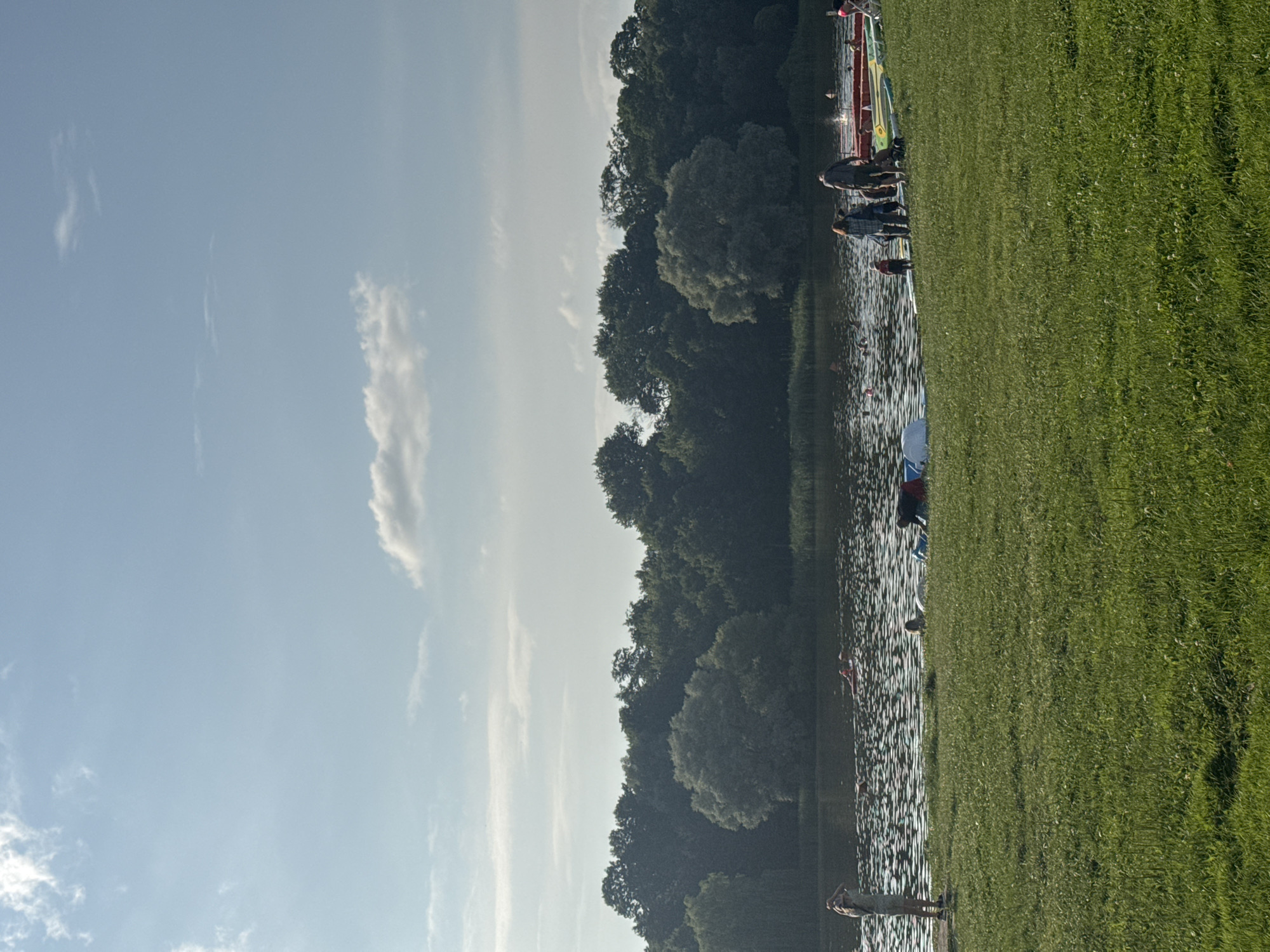 The image depicts a sunny day at a lake in Niasvizh, Belarus. The foreground is dominated by a lush green lawn with some scattered white flowers. In the midground, the lake is visible, shimmering with light reflections and dotted with small lily pads. Several people are present, some standing on the grassy bank and others in the water, engaged in various activities. One person is standing on the bank, facing away from the camera, with their arms raised. Further into the water, a person is visible on a red float. Near the edge of the water, a group of people are gathered, with some individuals appearing to be preparing to enter the water. Several colorful kayaks and paddleboards are lined up on the grassy bank next to the water. In the background, a dense forest of green trees forms a natural backdrop to the scene. The sky above is a clear blue with a few wispy white clouds.