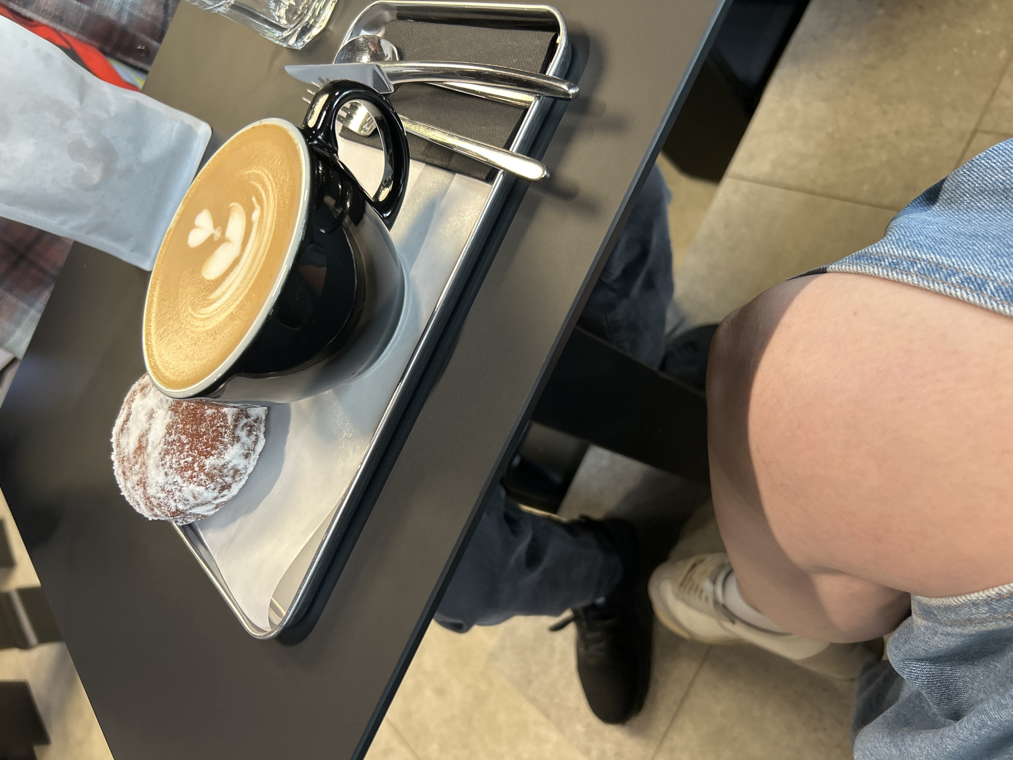 The image shows a tray on a black table with a black mug of latte and a powdered donut. There is a knife and fork on the tray, placed on a black napkin. A person is seated at the table, wearing jeans and a white sneaker on one foot and black sneaker on the other foot. The image does not provide enough detail to determine the specific location in Izhevsk, Russia.