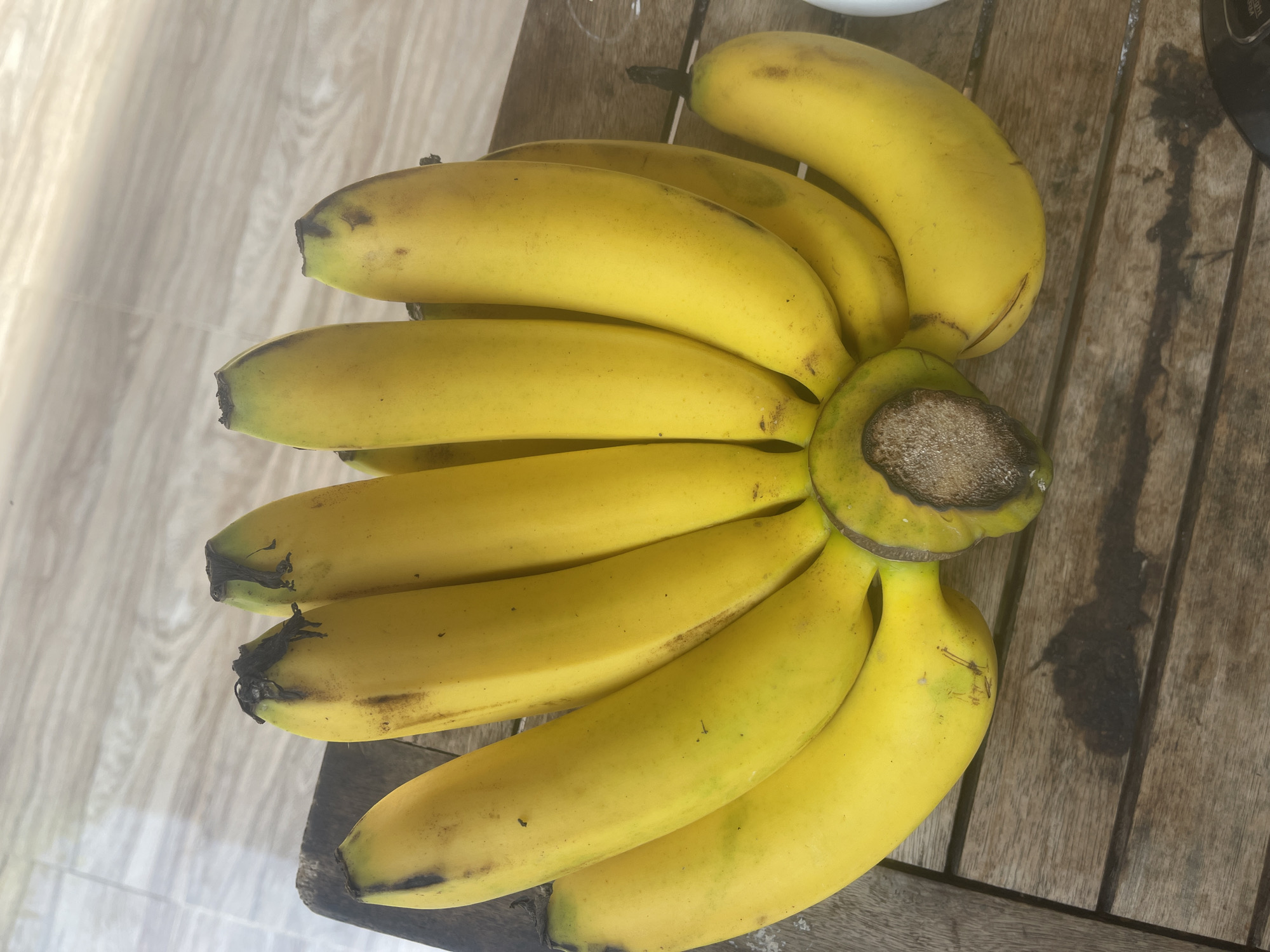 A bunch of yellow bananas lies on a wooden table. The bananas are ripe, with some brown spots on their peels. The table is made of weathered wooden planks, and the background shows a light-colored, textured surface, possibly a tiled floor, with soft shadows cast by light sources from above. The scene appears to be outdoors, given the natural lighting and the outdoor setting implied by the materials. No people are visible in the image. The image is shot from a top-down perspective.