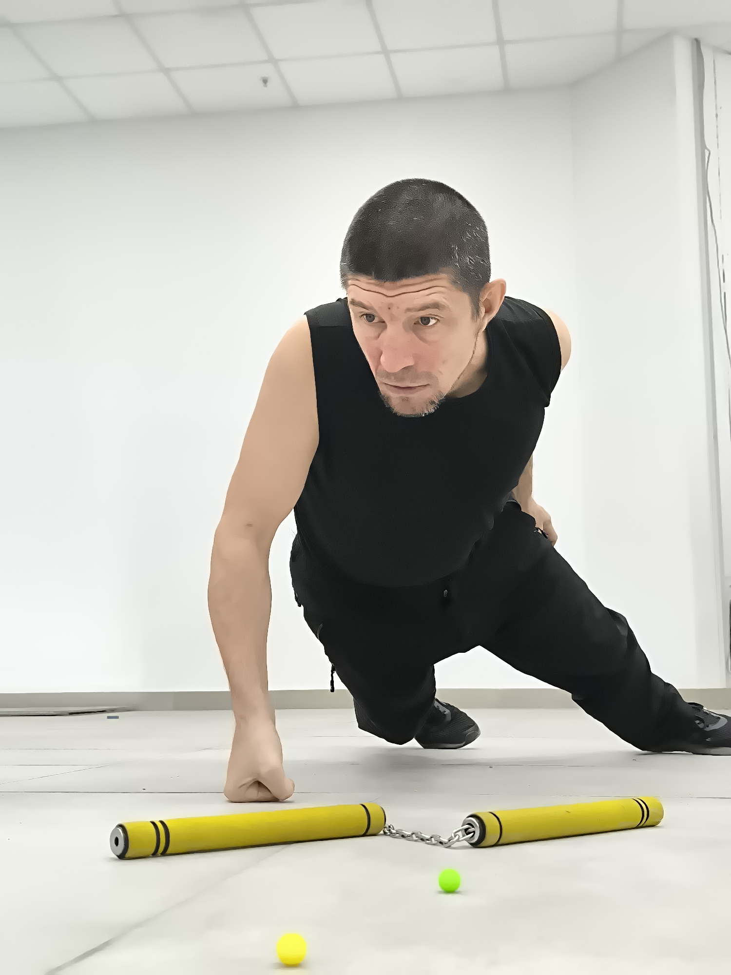 A man in athletic wear is in a push-up position on a tiled floor. He is wearing a black sleeveless shirt and black pants. His body is angled towards the right of the frame, and his head is turned to look to his left. In front of him on the floor are two yellow nunchucks connected by a silver chain. There is also a small green ball and a small yellow ball on the floor, spaced between the nunchucks. The background is a plain white wall and ceiling with fluorescent lighting. The location is not specified as being in the city of Qarağandı city administration, Kazakhstan.