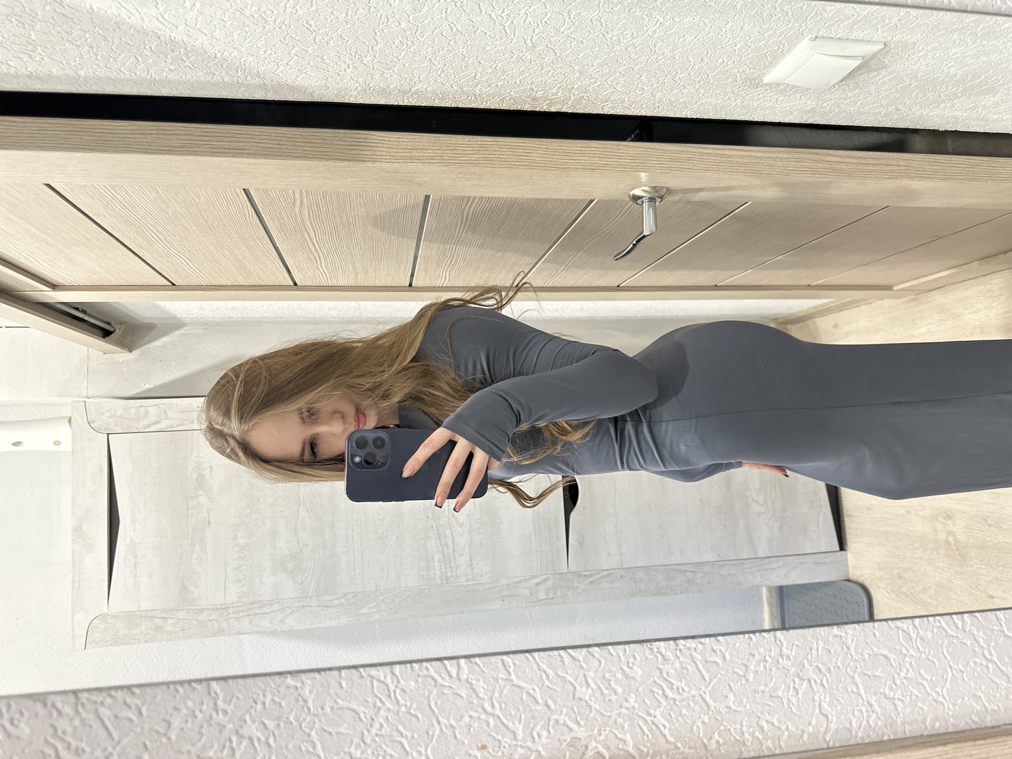 The image is a selfie taken in a mirror. A young woman with long brown hair is holding a dark blue smartphone, angled so that her face is visible in the reflection. She is wearing a long-sleeved, grey bodycon dress. The mirror reflects a room with a light-colored wooden door, white walls with textured wallpaper, and white cabinets. The location is Unknown, Unknown.