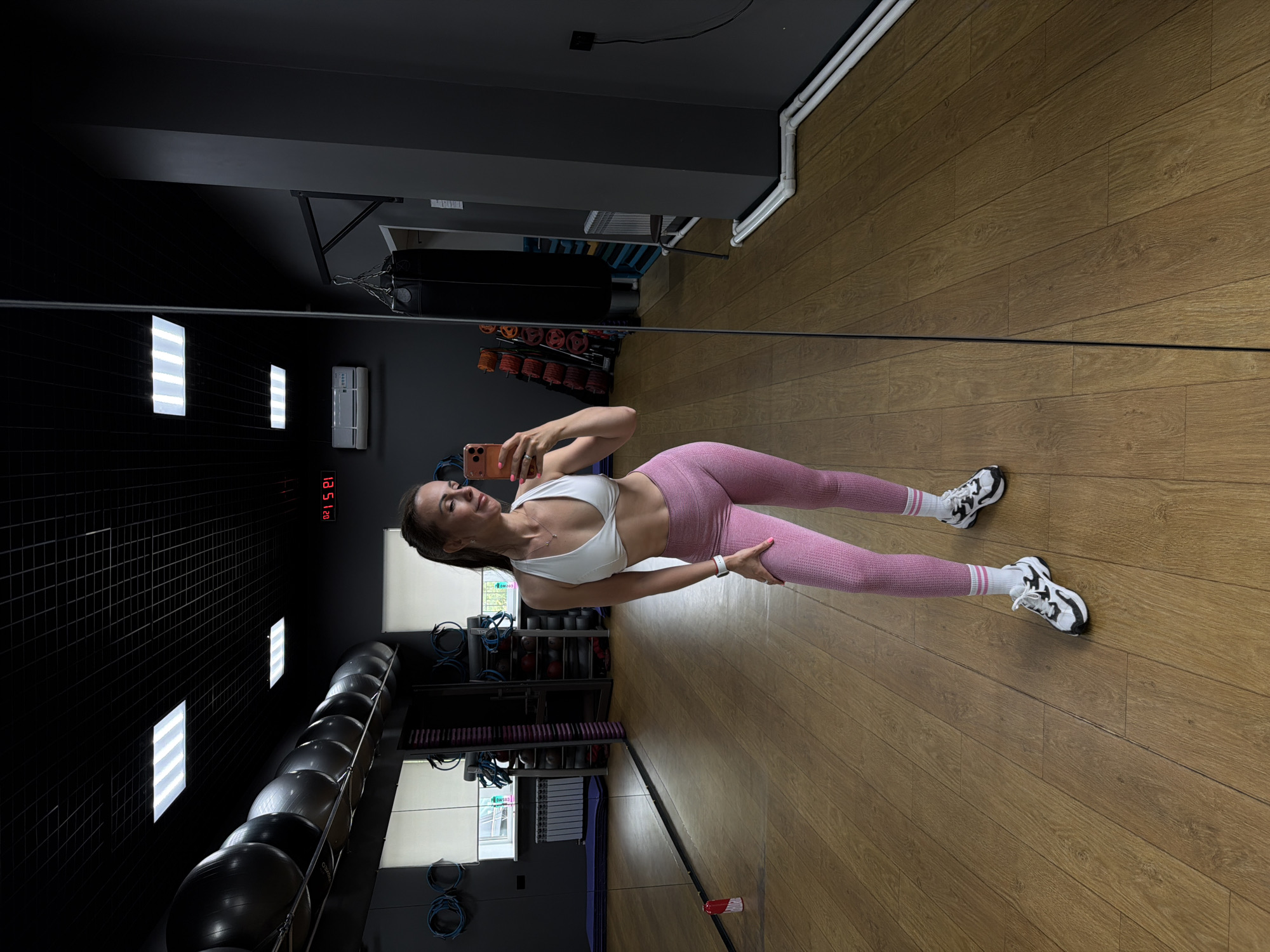 A woman is taking a mirror selfie in a gym in Birobidzhan, Russia. She is wearing a white sports bra and pink leggings, with white socks and black and white athletic shoes. She is holding a smartphone in her right hand, and her left hand is on her hip. The gym has a wooden floor, large mirrors, and various exercise equipment, including a punching bag, exercise balls, and weights. The room is lit by fluorescent ceiling lights. The time on a digital clock in the background reads 21:52.