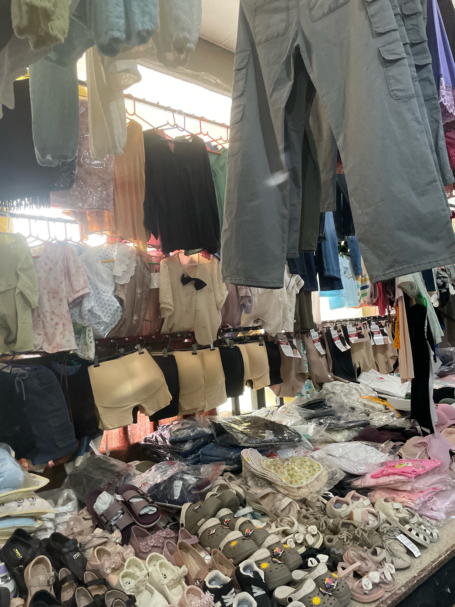 The image depicts an outdoor market stall in Kangpokpi, India, displaying a variety of clothing and footwear for sale. In the foreground, a large assortment of children's shoes, including sandals and clogs, are arranged on a surface. Above this, various undergarments, such as bras and shapewear, are hung on display. Further back, a rack holds an array of shirts and pants, some of which appear to be cargo pants. The stall is covered by a makeshift roof, likely made of tarpaulin or similar material, and a bright light source is visible above, suggesting it might be evening or a dimly lit area. The overall scene is one of a busy marketplace with items neatly but densely displayed. There are no people visible in the frame.