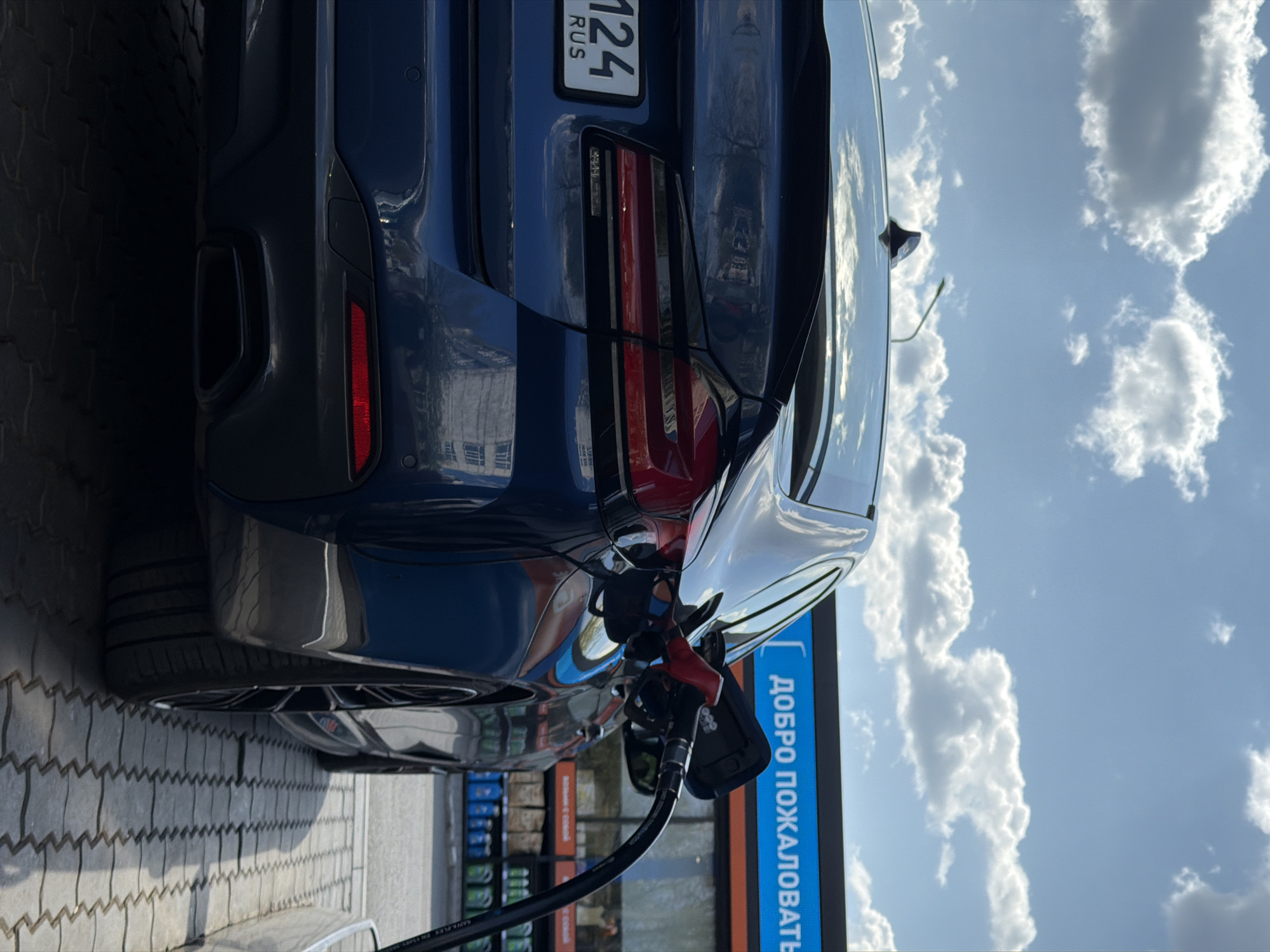 The image shows the rear of a dark-colored car being refueled at a gas station. The car has a license plate with "124" and "RUS" visible, indicating registration in Russia. The gas pump is connected to the car. In the background, a sign reads "ДОБРО ПОЖАЛОВАТЬ" (Welcome) in Russian. The sky is visible in the background, with white clouds against a blue sky. The image was likely taken at a gas station in Krasnoyarsk, Russia. The time of day appears to be daytime due to the clear sky.