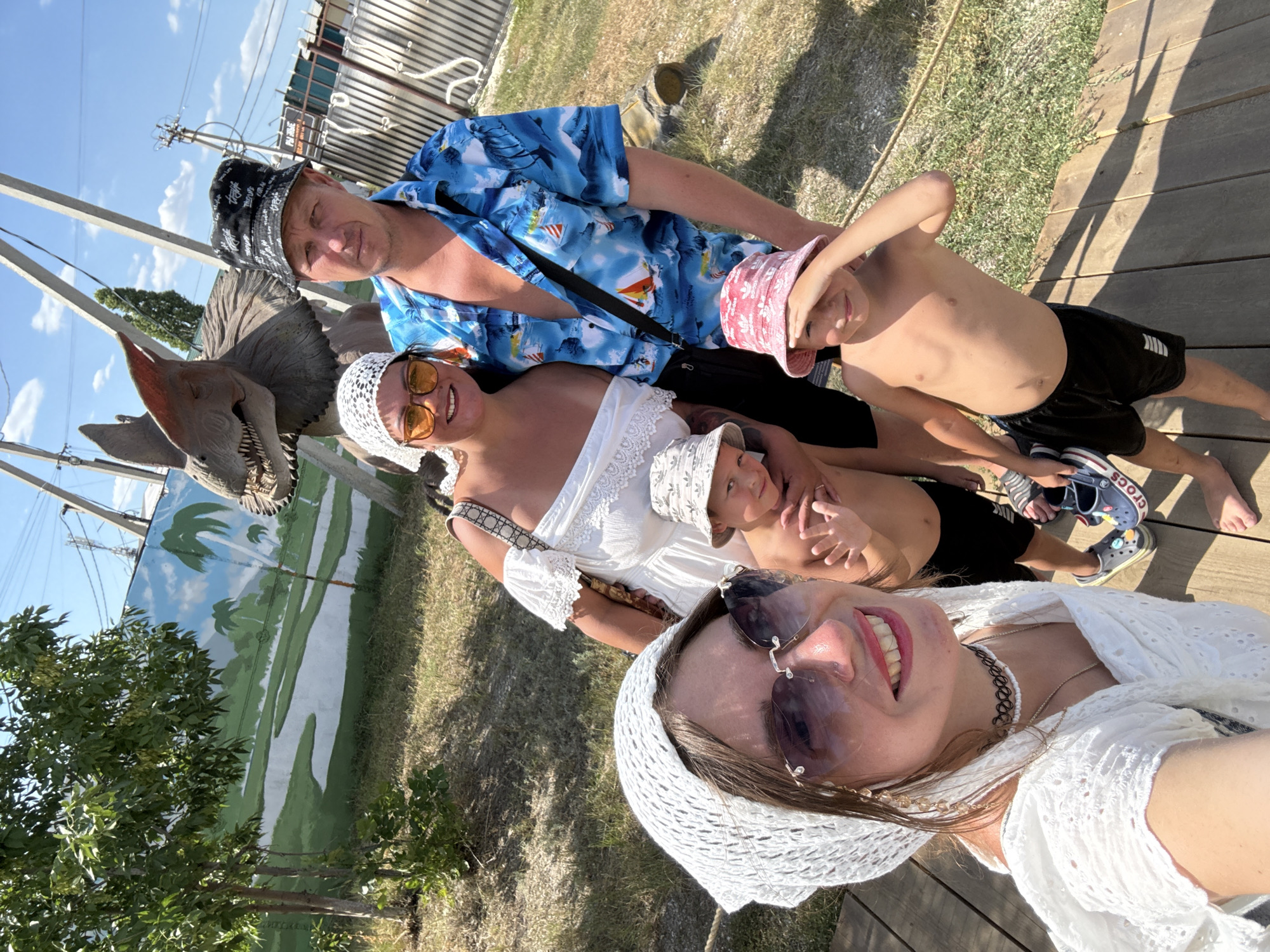 In Sol-Iletsk, Russia, a family of four poses for a selfie outdoors on a sunny day. The group includes two adults and two young boys. The woman in the foreground, wearing a white headscarf and sunglasses, is holding the camera. To her right, an adult male wearing a blue Hawaiian shirt and a bucket hat stands next to another woman with orange-tinted sunglasses and a white headscarf. The two boys are positioned in front of the adults. One boy, wearing a bucket hat, is being held by the woman, while the other boy, also wearing a bucket hat, stands next to him. Behind the family, a statue of a dinosaur with a frill around its neck is visible, along with a mural depicting a prehistoric landscape with palm trees. The ground is covered in wooden planks and dry grass.