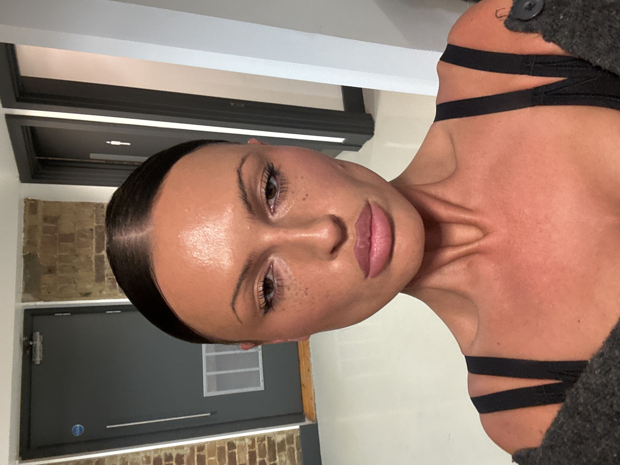 A close-up, upright portrait captures a person, likely a woman, with luminous skin and subtly applied makeup. Her dark hair is slicked back tightly, emphasizing her sculpted features, including defined eyebrows, long eyelashes, and lips with a soft pink or nude gloss. Her gaze is direct and composed.
She is wearing a black top with multiple thin, horizontal straps visible across her right shoulder and collarbone area, suggesting a contemporary design. A dark, textured garment, possibly a grey sweater or jacket, is worn over it, with a black button visible near her right shoulder.
The background is an indoor setting with a modern, minimalist aesthetic. To the viewer's left, a dark grey door with a silver handle is partially visible, with hints of exposed brickwork near the bottom. To the viewer's right, a light-colored wall frames the subject. Above the door frames, linear light fixtures or ceiling details are present. The lighting is bright and even, highlighting the person's features. There is a small circular blue sticker on the dark grey door. No visible text is present in the image.