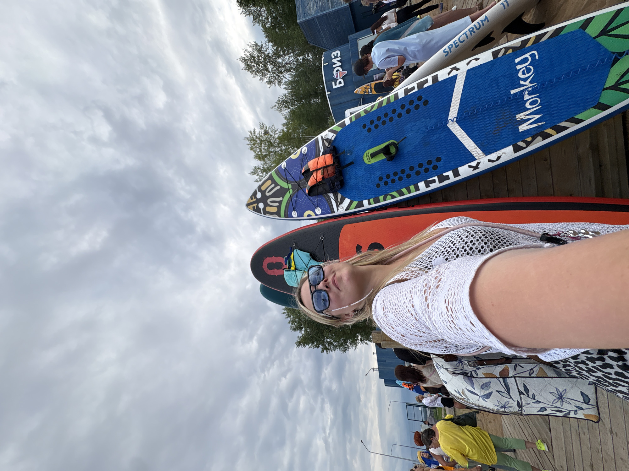 A group of people are gathered outdoors on a wooden platform near a body of water. Several stand-up paddleboards (SUPs) are laid out, some with colorful designs. In the foreground, a woman with blonde hair, wearing sunglasses and a white mesh top, is taking a selfie. She is leaning back, with the SUPs behind her. Other individuals are visible in the background, some standing, some sitting.
One SUP board has the word "Monkey" written on it in white lettering. Another has "SPECTRUM" written on it. A building with the Cyrillic word "БИЗ" is visible in the background. The sky is overcast with gray clouds, suggesting a cloudy day. The scene appears to be a recreational area, possibly a beach or a waterfront park, given the presence of SUPs and the wooden decking. The overall mood seems relaxed and casual. The time of day is likely daytime.